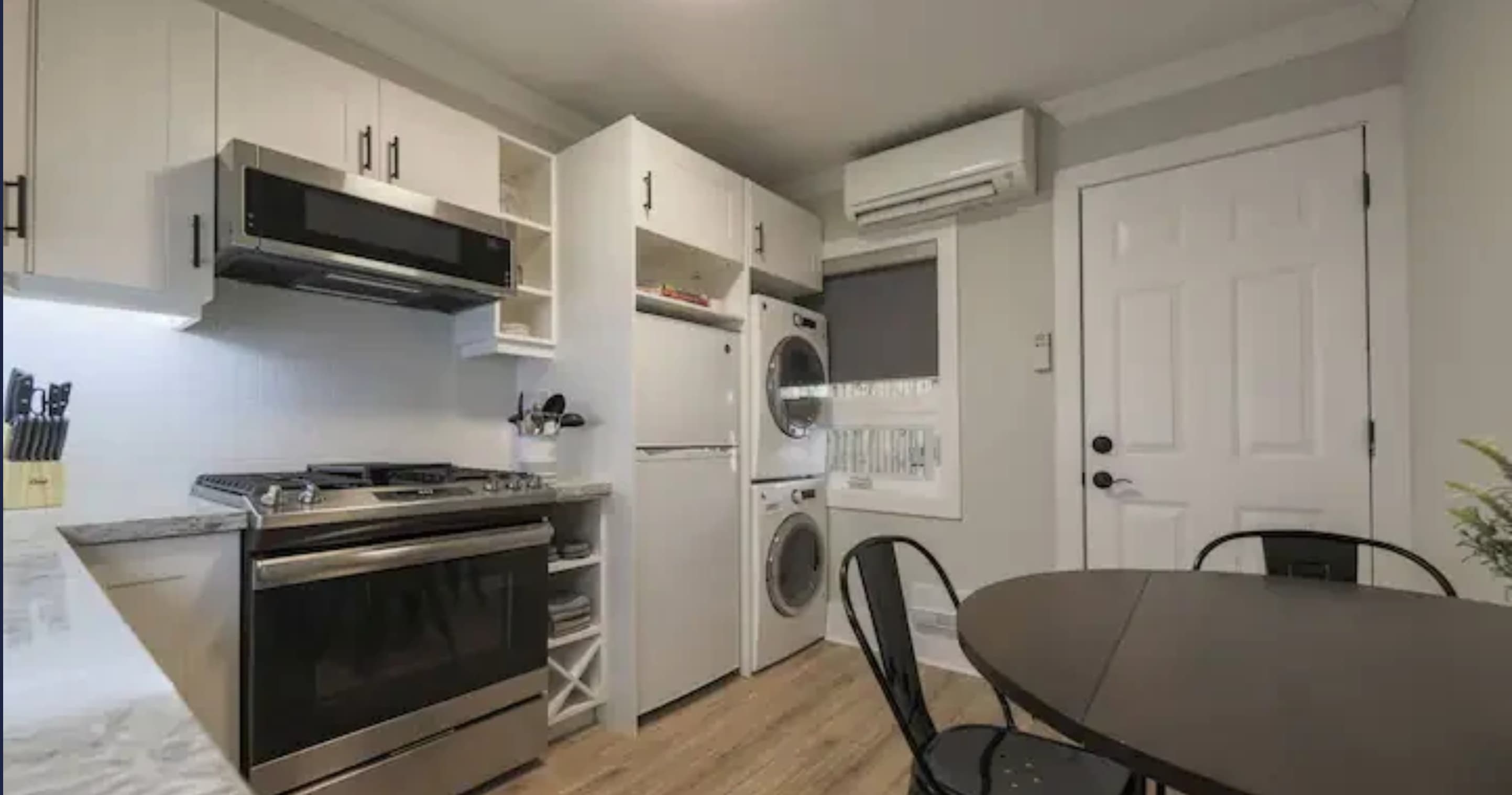 Full view of the modern kitchen with white cabinets, stainless steel appliances including a gas range, microwave, and refrigerator. A stacked washer and dryer are visible next to the fridge, and a small dining table is in the foreground.