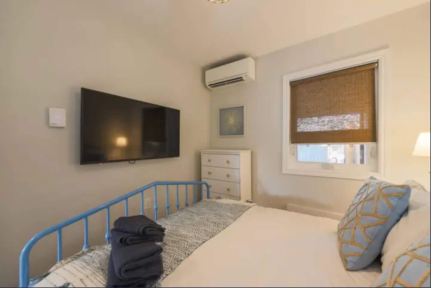 Cozy bedroom featuring a blue metal-frame bed with white and blue bedding, a wall-mounted TV, and a white dresser. A window with a woven blind provides natural light, complemented by an AC unit mounted above.
