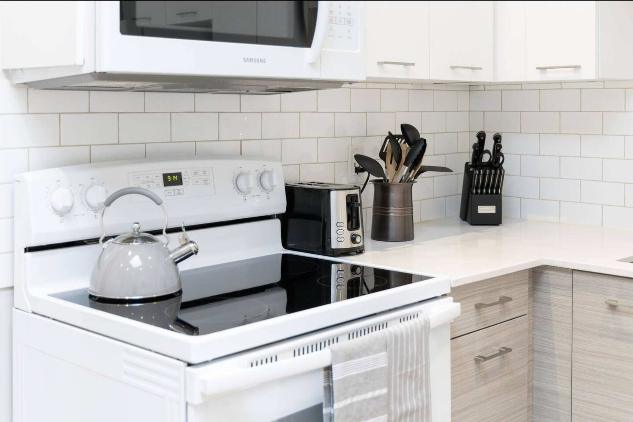 Close-up view of the kitchen countertop and white electric stove, featuring a subway tile backsplash, a grey kettle, toaster, and various cooking utensils in a bronze holder.