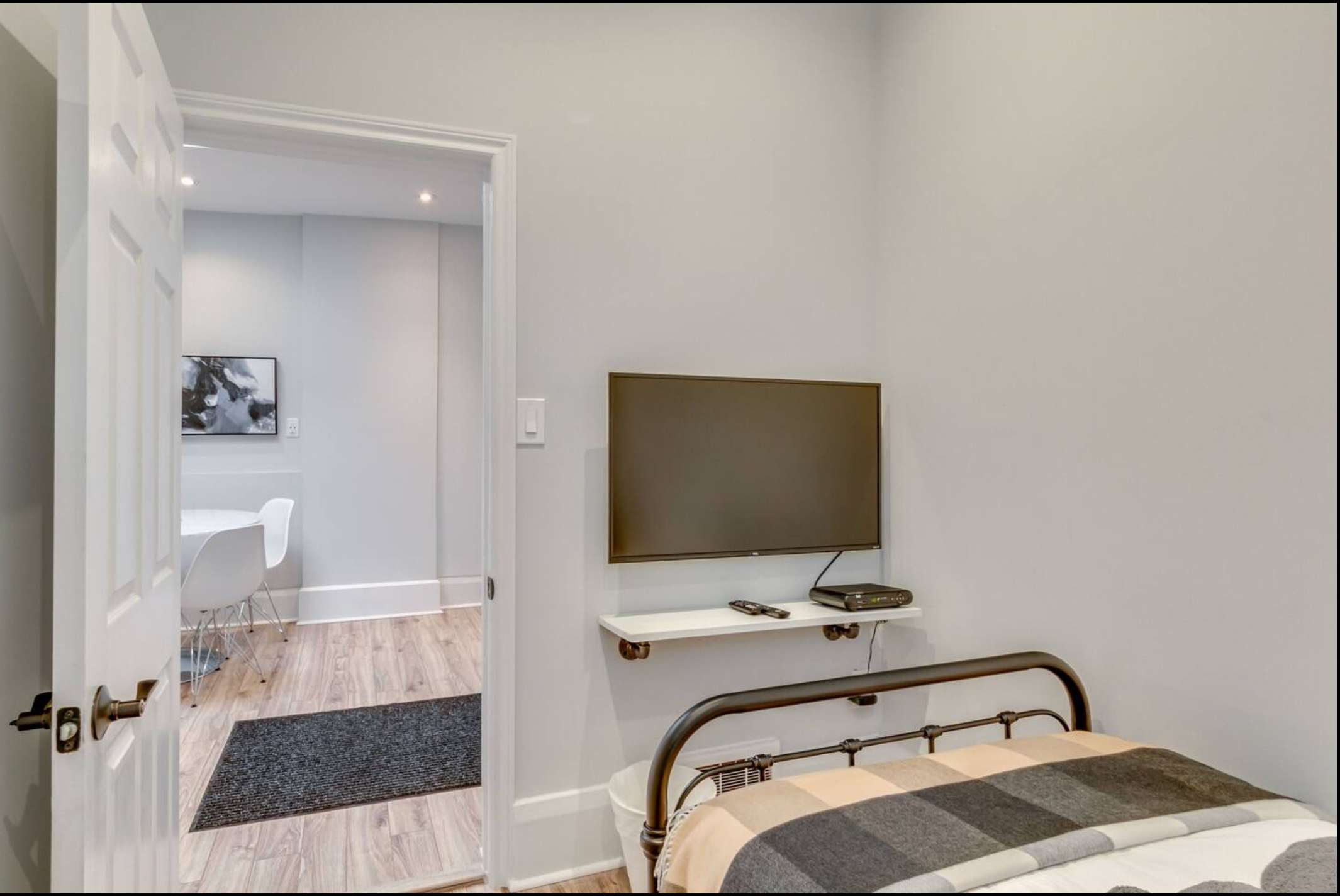 View from a bedroom with a wall-mounted TV and a black metal bed frame, looking through an open doorway into an adjacent room with a white dining table and chairs.
