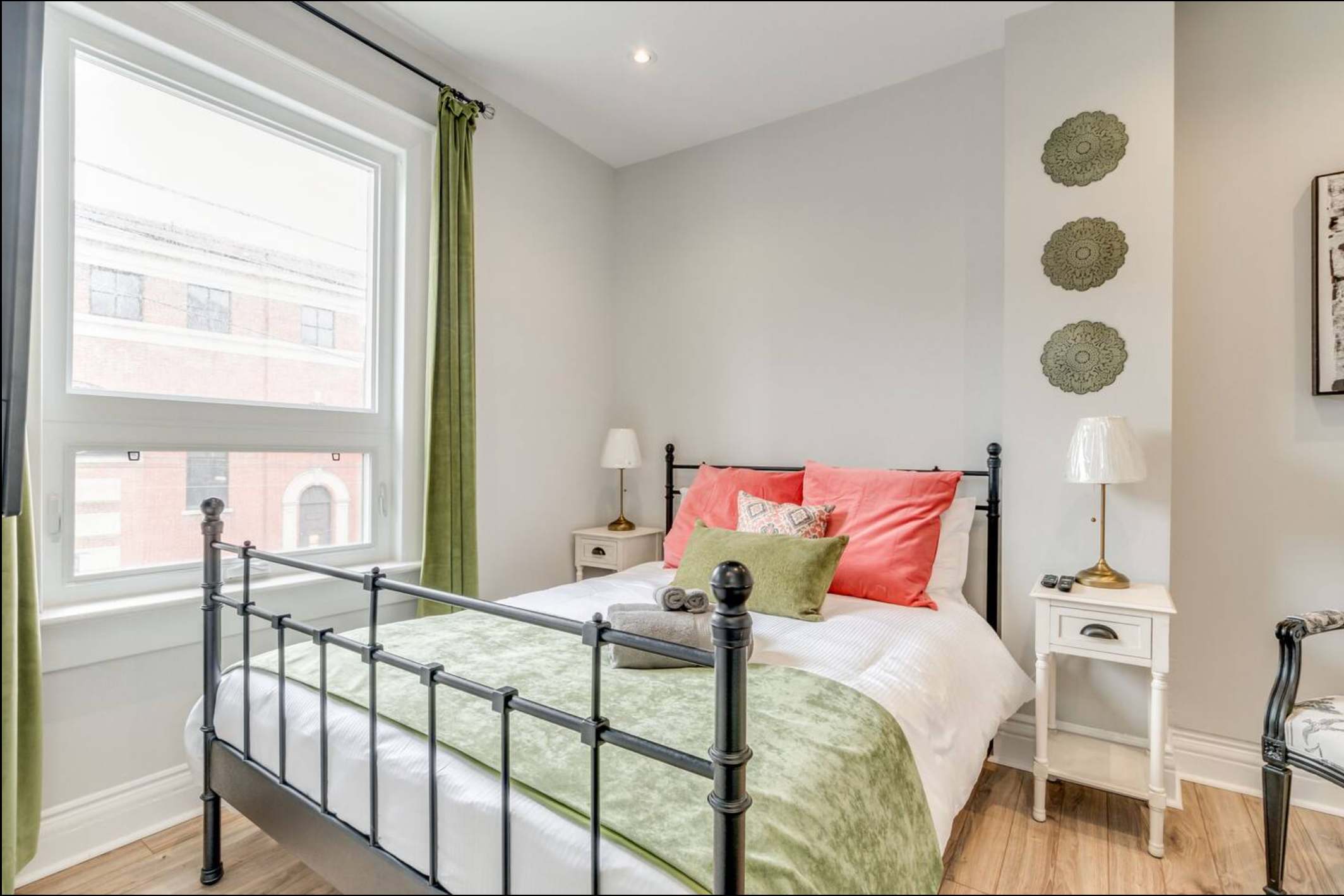 Cozy bedroom with a black metal bed frame, white and green bedding, coral pillows, and two white nightstands with lamps, positioned beneath a large window with green curtains. Decorative wall art is visible on the right.