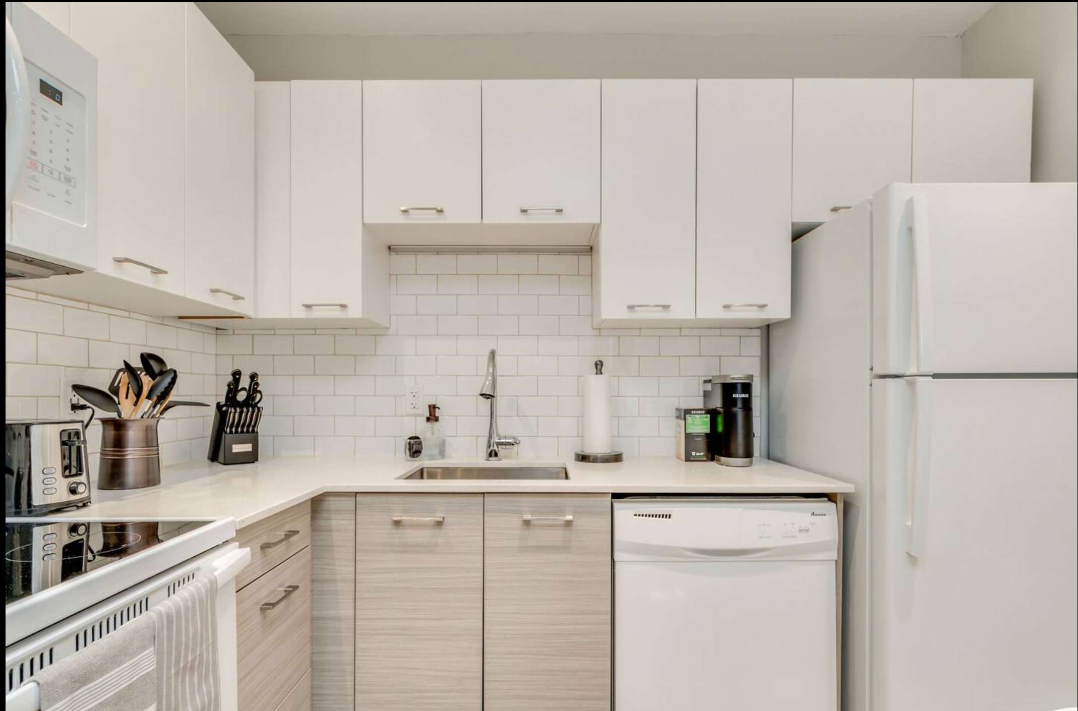 Detailed shot of the kitchen showing the stainless steel sink, white dishwasher, and light wood-look lower cabinets contrasting with white upper cabinets and a clean subway tile backsplash.