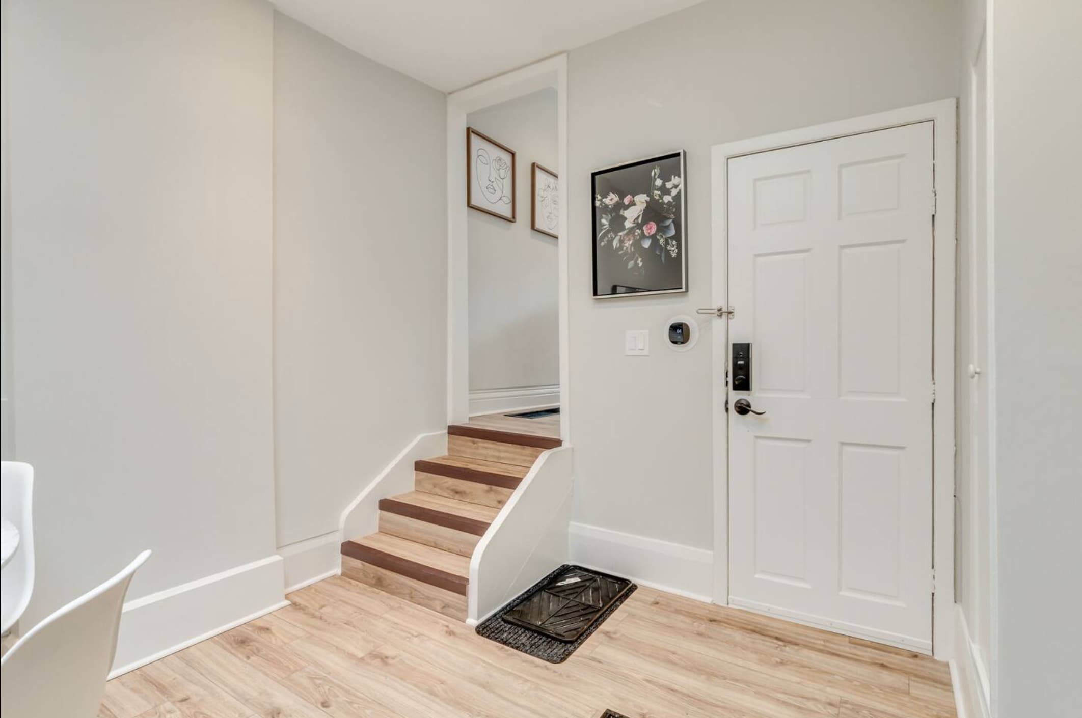 Bright entryway featuring light hardwood floors, a short set of wooden stairs leading up, and a white front door with a smart lock. Decorative framed art adorns the wall.