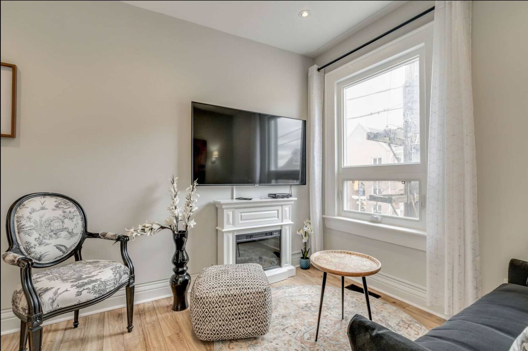 Another view of the living room, showcasing a wall-mounted flat-screen TV above a white decorative electric fireplace, an ornate accent chair, and a large window with sheer white curtains.