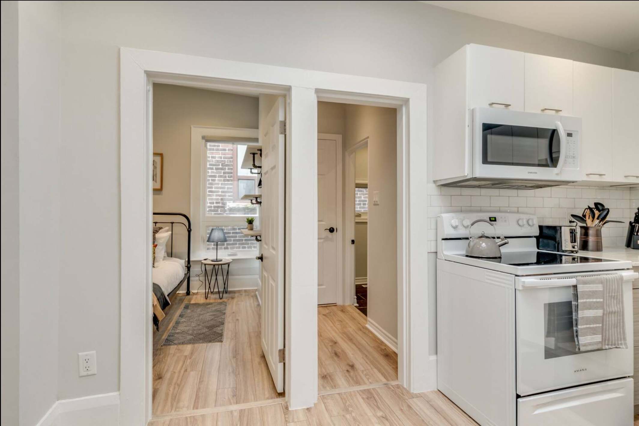 Modern kitchen with white cabinetry, subway tile backsplash, and a white electric stove, offering a glimpse into a bedroom with a black metal bed frame and a window.