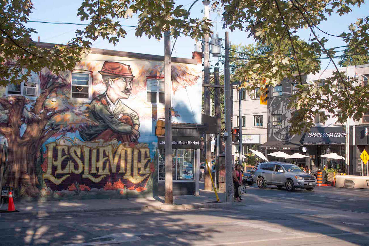 Vibrant street scene in Leslieville featuring a large colorful mural of a man in a hat on a brick building, with cars and pedestrians on the street, surrounded by green trees.
