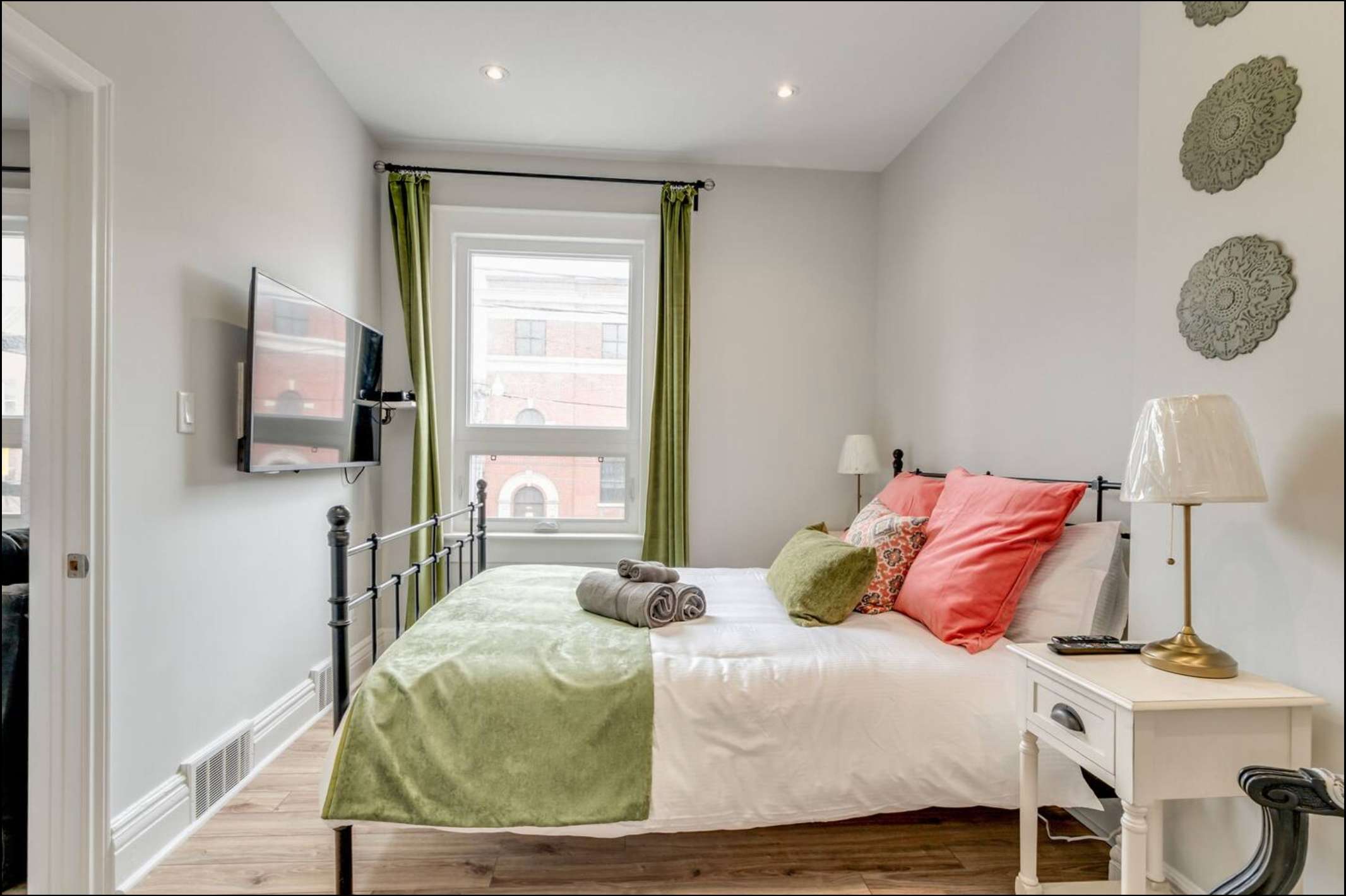 Bright bedroom featuring a black metal bed frame with white and green bedding, coral pillows, two white nightstands with lamps, and a wall-mounted TV opposite a large window with green curtains.