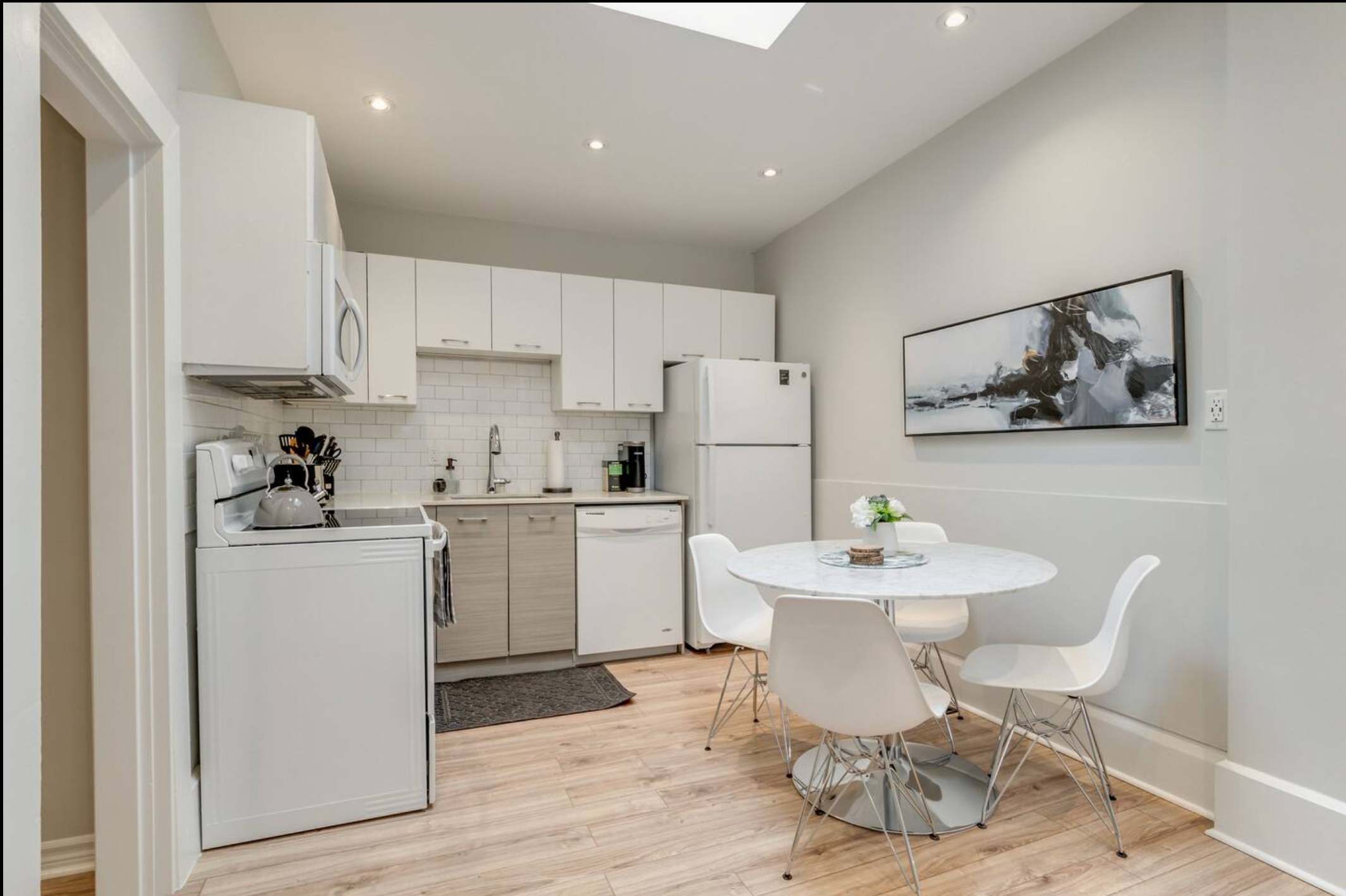 Spacious kitchen and dining area with light wood-look flooring, white cabinetry, a skylight, and a round white dining table with four modern white chairs, creating an airy atmosphere.