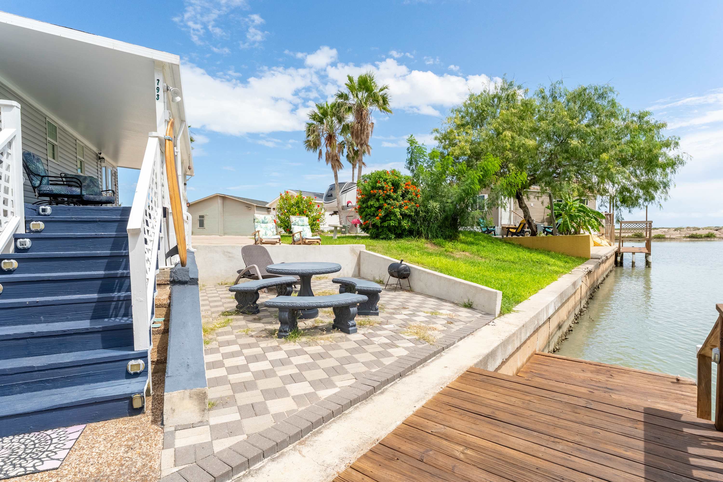An outdoor patio area with a checkered paver floor, a stone table and benches, and a grassy yard leading to a canal, surrounded by lush greenery and palm trees, with a BBQ grill.