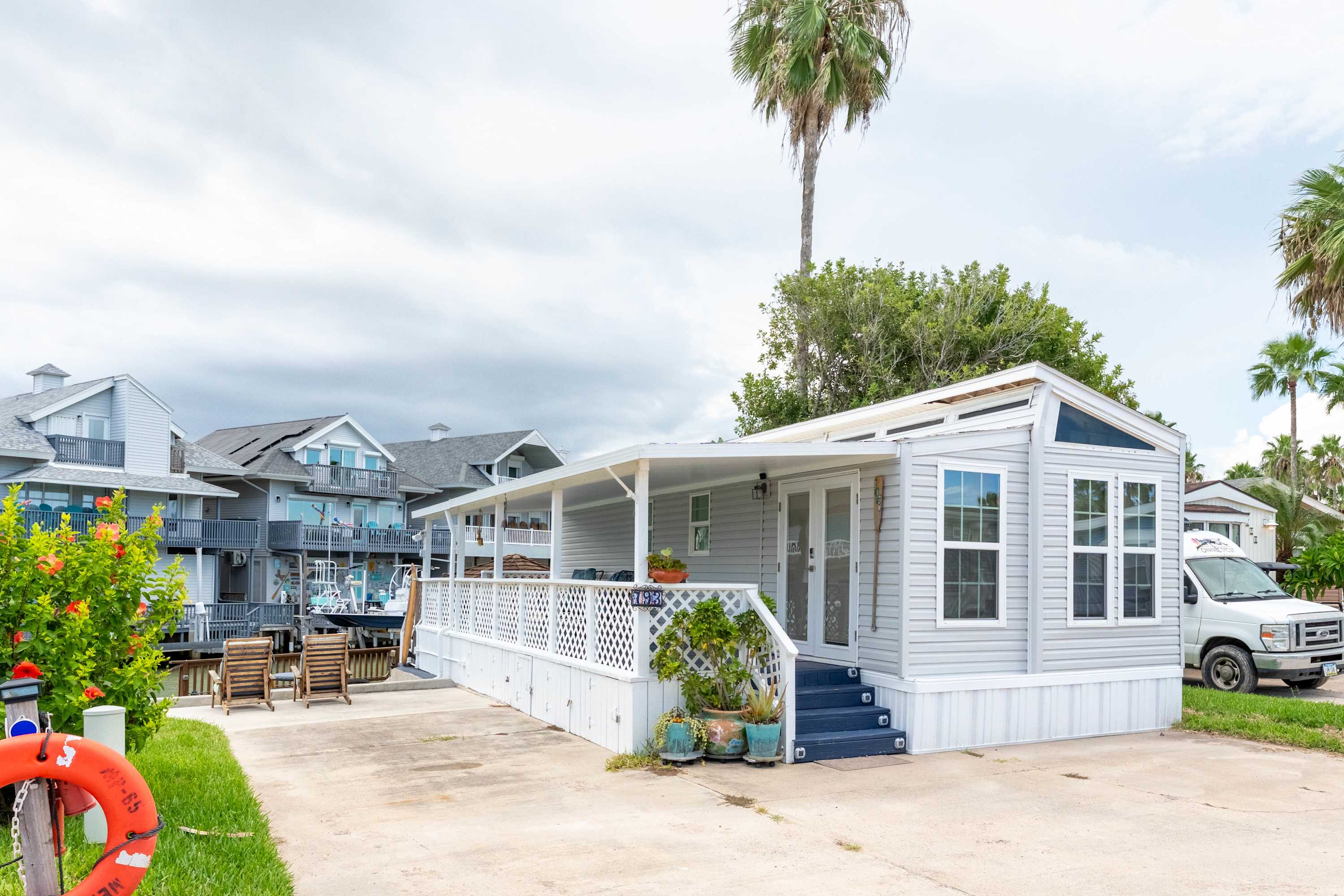 The front exterior of a light gray mobile home with white trim, a covered porch, and blue steps, situated next to a paved driveway and a tall palm tree, with a view of the canal in the background.