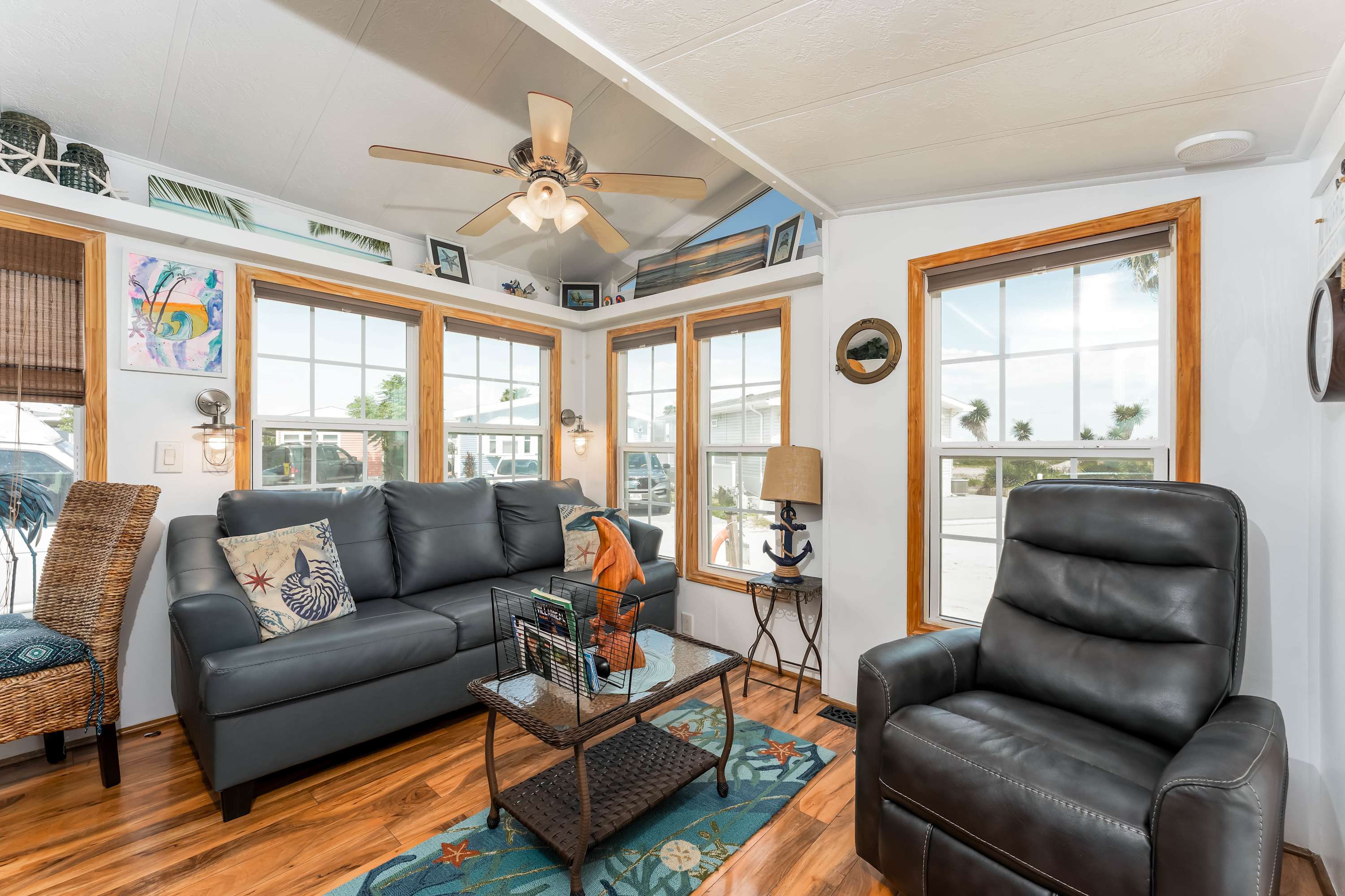 A bright living room with wood-look flooring, a dark gray sofa, a matching recliner, and multiple windows with wood trim, featuring a ceiling fan and coastal decor.