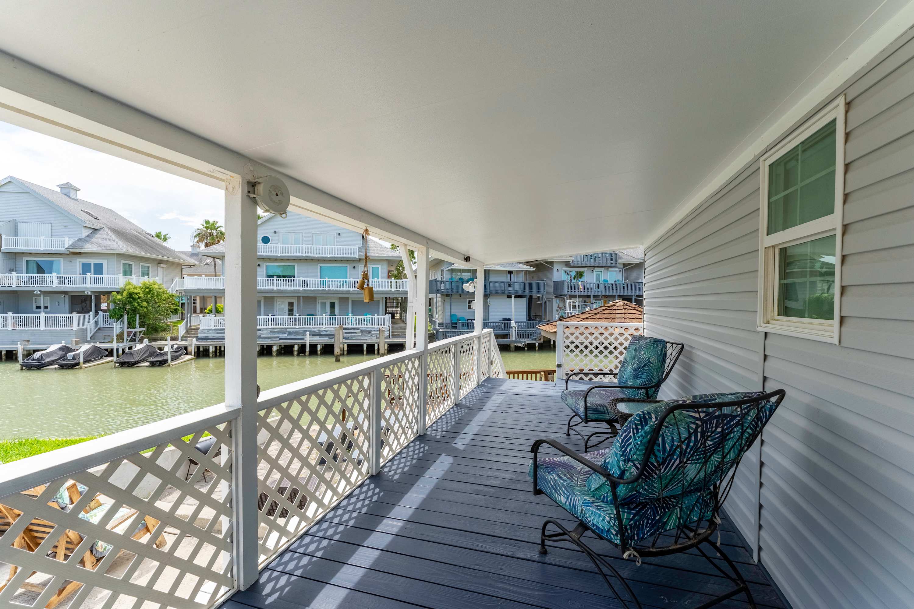 A long, covered outdoor deck with dark blue flooring and white lattice railings, furnished with two metal chairs and a side table, offering a view of the canal and neighboring properties.