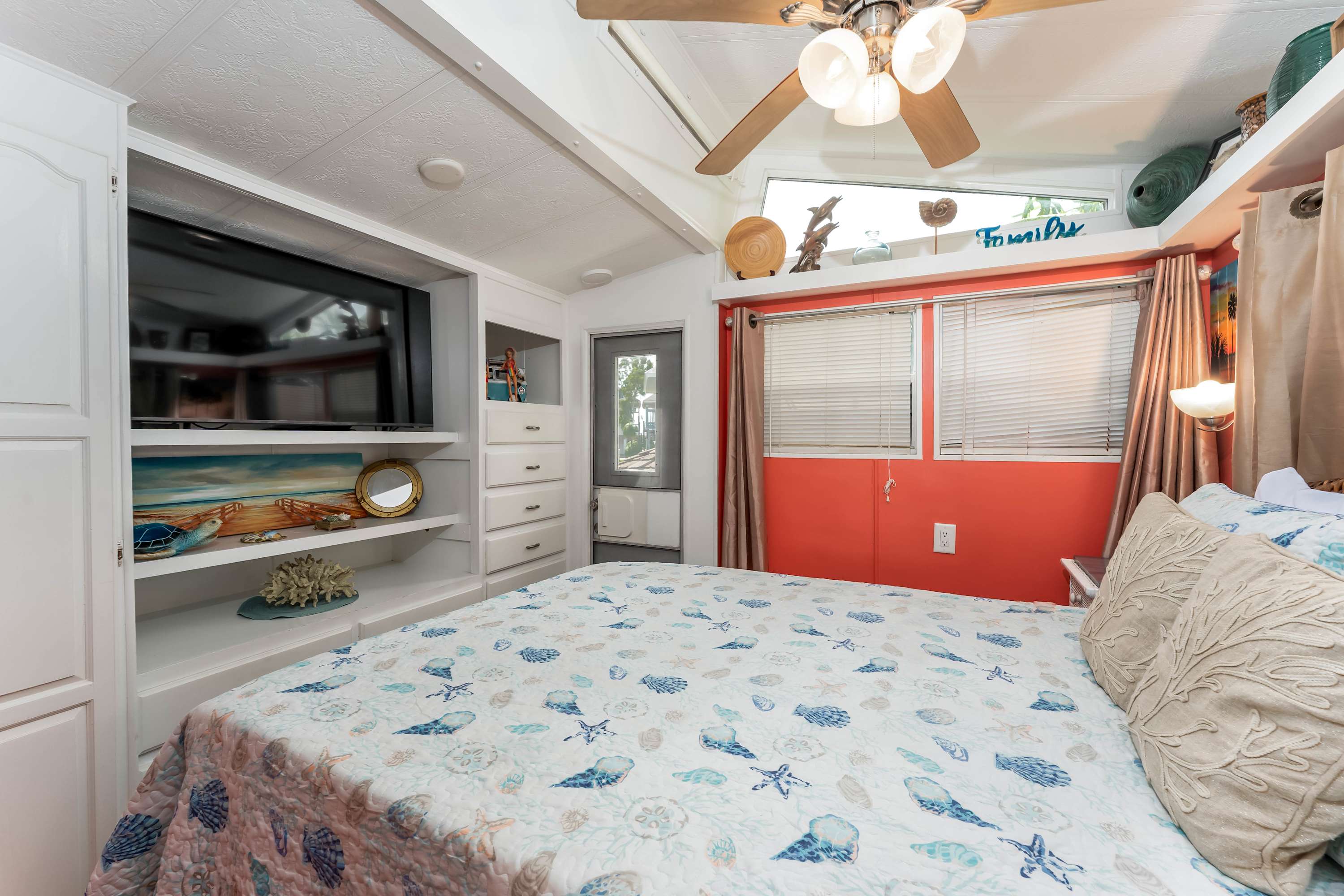 A bright bedroom with a bed covered in a white and blue seashell-patterned quilt, built-in white cabinetry with a TV, and a window with orange blinds, featuring a sloped ceiling and ceiling fan.