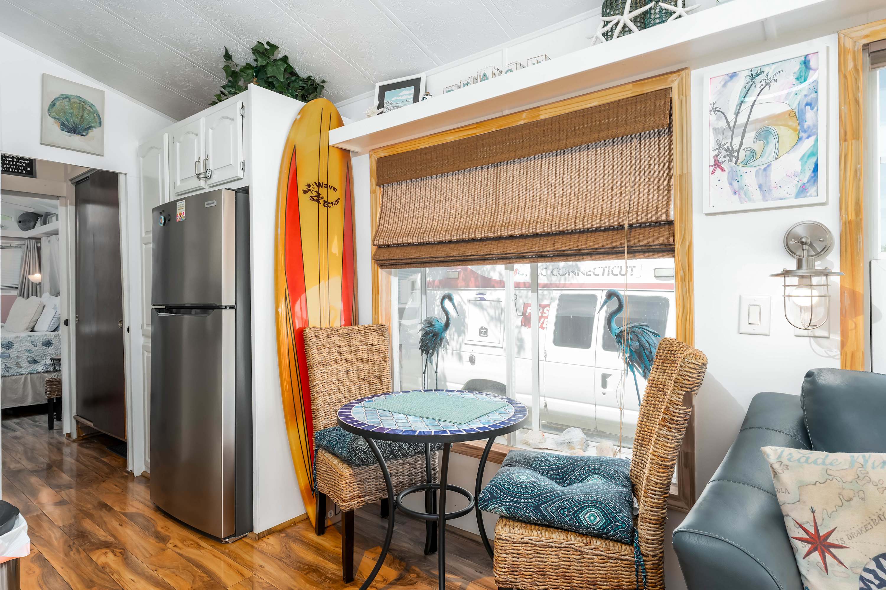 A corner of the living room with wood-look flooring, a small round dining table with two wicker chairs, and a large window with a bamboo blind, featuring a tall surfboard and a refrigerator.