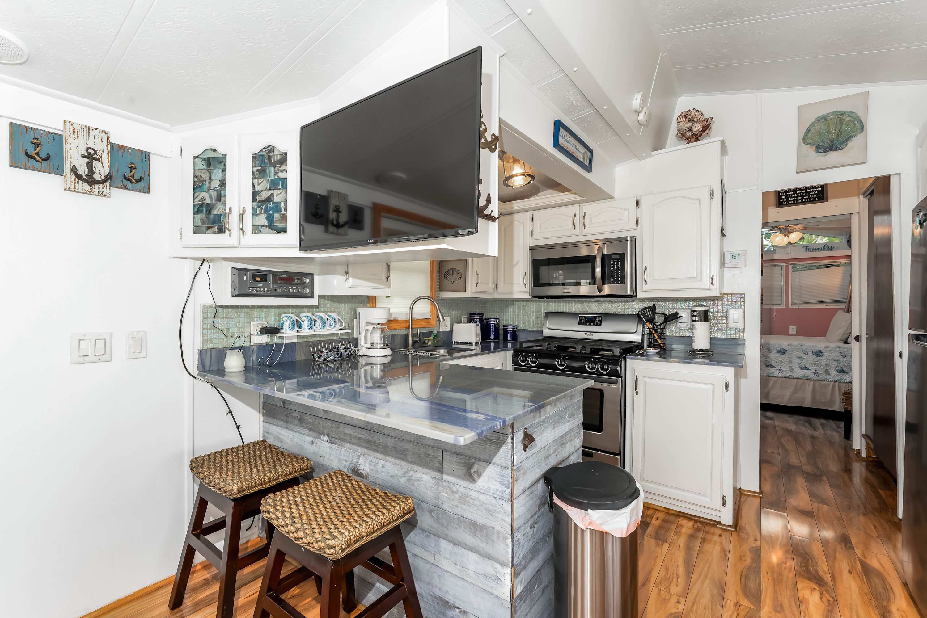 A compact kitchen with white cabinets, stainless steel appliances, a blue-gray countertop, and a breakfast bar with two woven stools, featuring a wall-mounted TV and wood-look flooring.