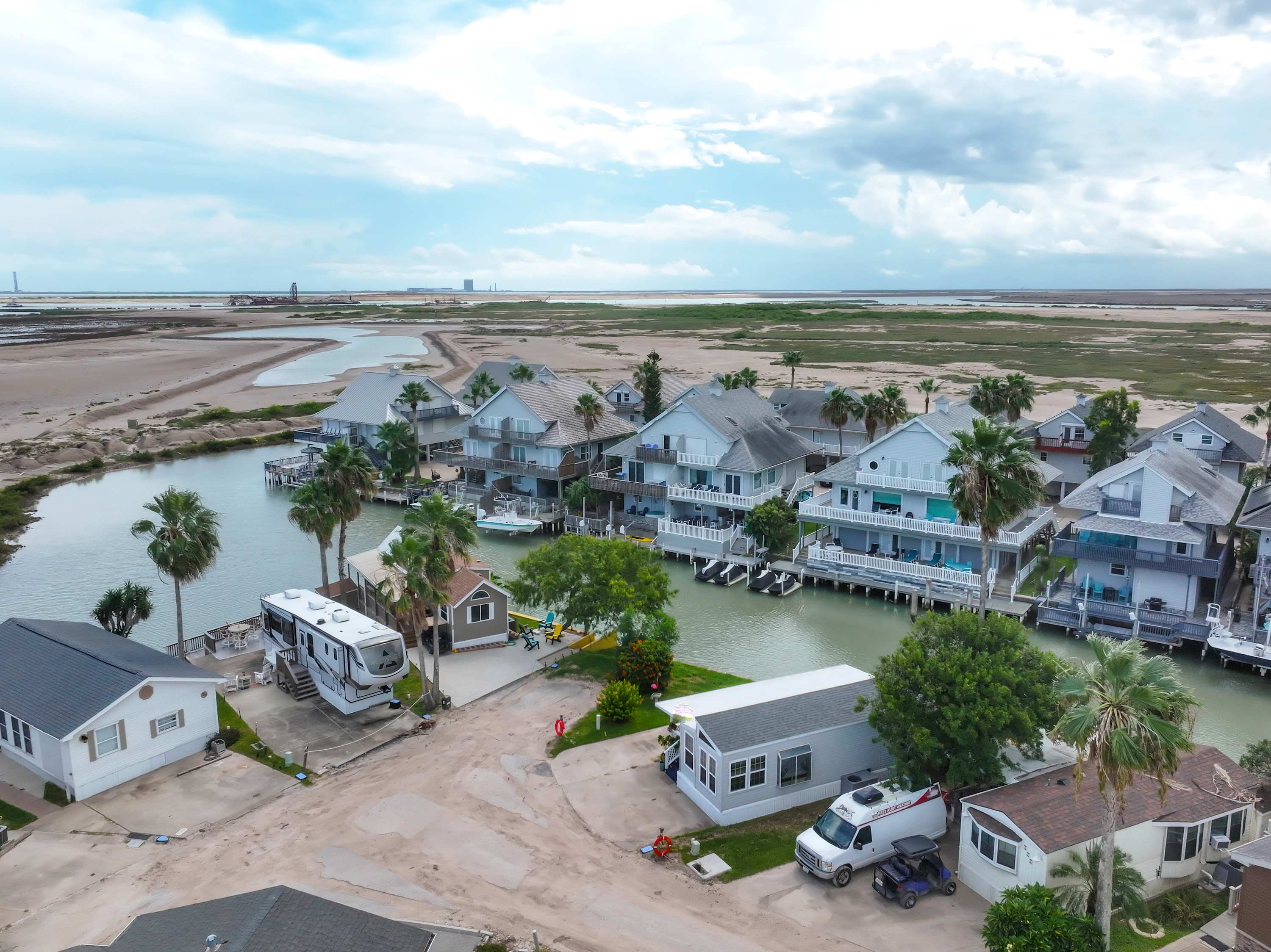 An expansive aerial view of a vibrant waterfront community, showcasing numerous houses with boat slips lining intricate canals. The landscape is dotted with palm trees, leading to a distant bay and industrial structures under a dynamic sky.