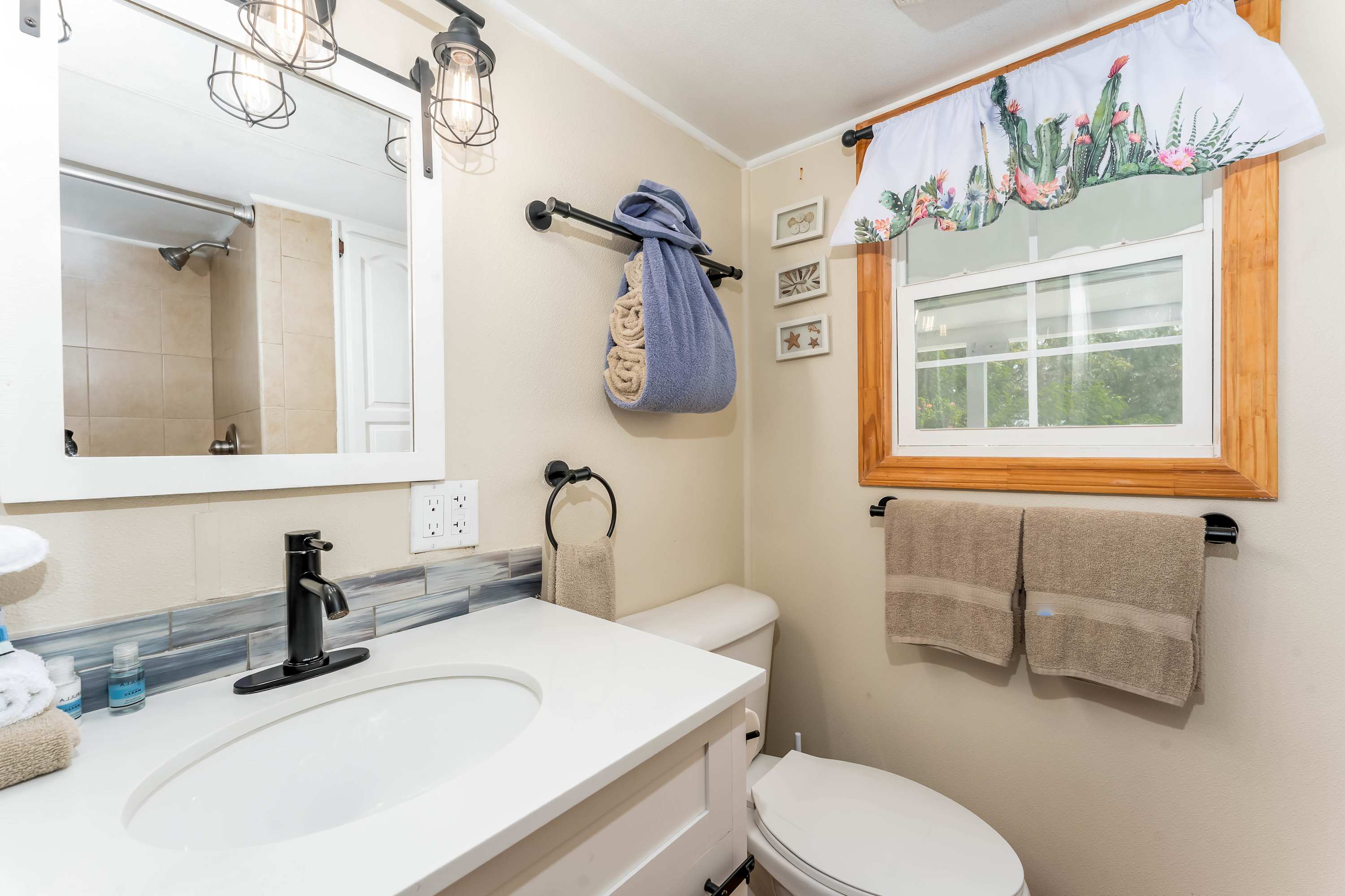 A modern bathroom with a white vanity and sink, black faucet, a rectangular mirror with industrial-style lighting, and a window with a cactus-patterned valance, featuring a blue tiled backsplash.