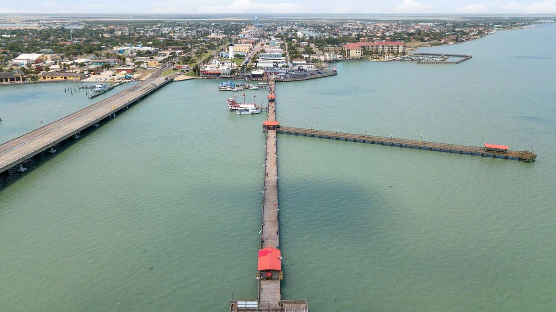 An impressive aerial view of a very long wooden pier extending far into a wide body of water, with a parallel bridge connecting to a bustling coastal town in the background. The water is calm under a bright, partly cloudy sky.