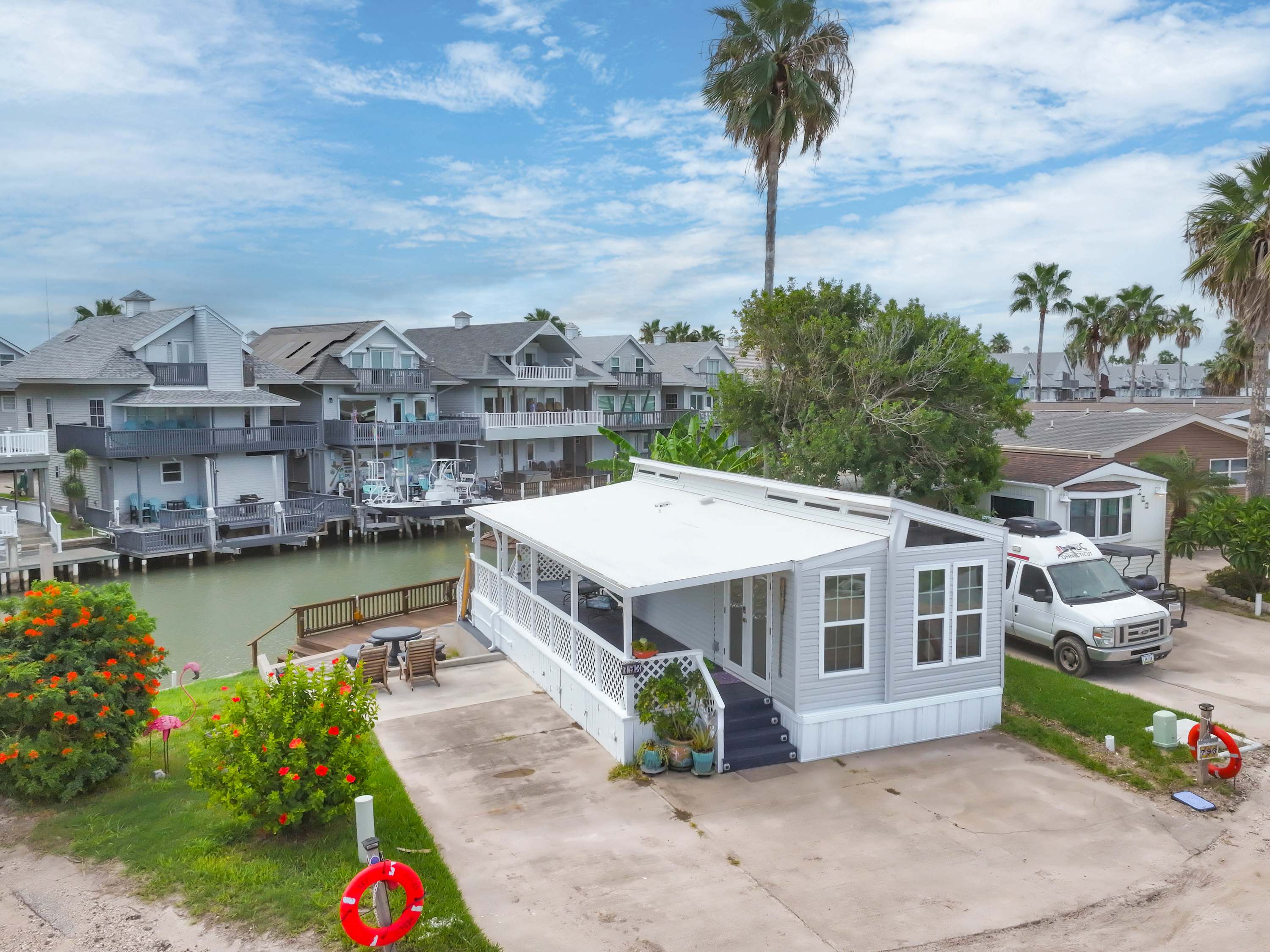 A charming light gray mobile home or small house with white trim and a spacious covered front porch, featuring outdoor seating. It's situated by a canal with a small dock, surrounded by lush green landscaping and palm trees.