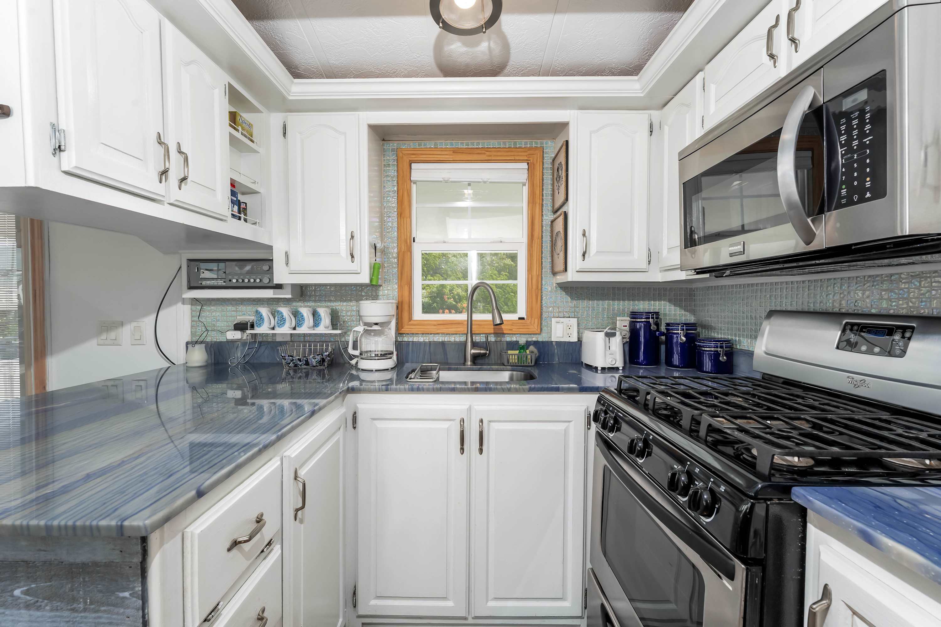 A bright kitchen with white shaker cabinets, blue marble-patterned countertops, a blue tiled backsplash, and stainless steel appliances, featuring a window above the sink.