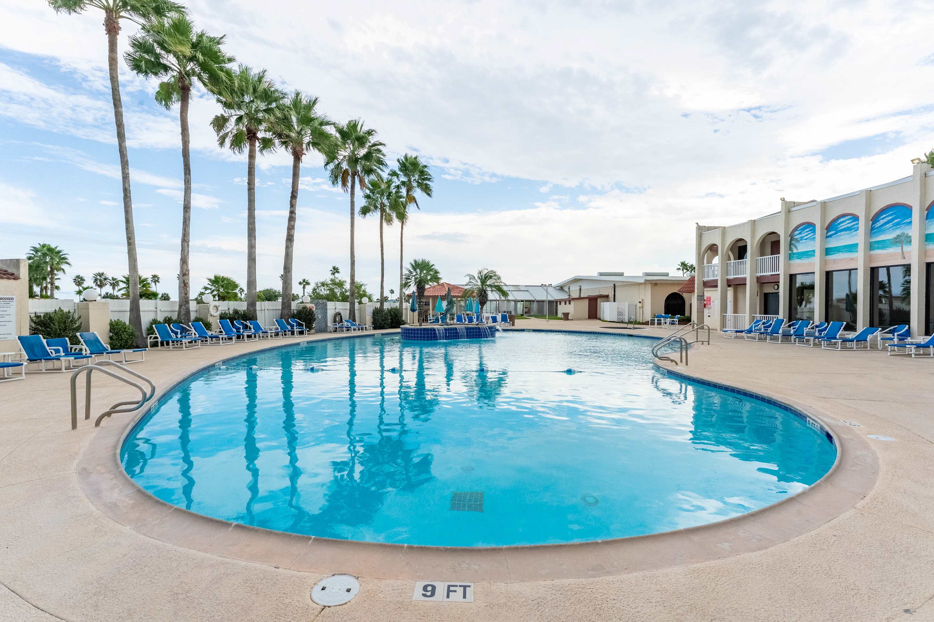 A large, inviting kidney-shaped outdoor swimming pool with clear blue water, surrounded by a concrete deck with blue lounge chairs. Tall palm trees and a multi-story building with arched windows frame the background.