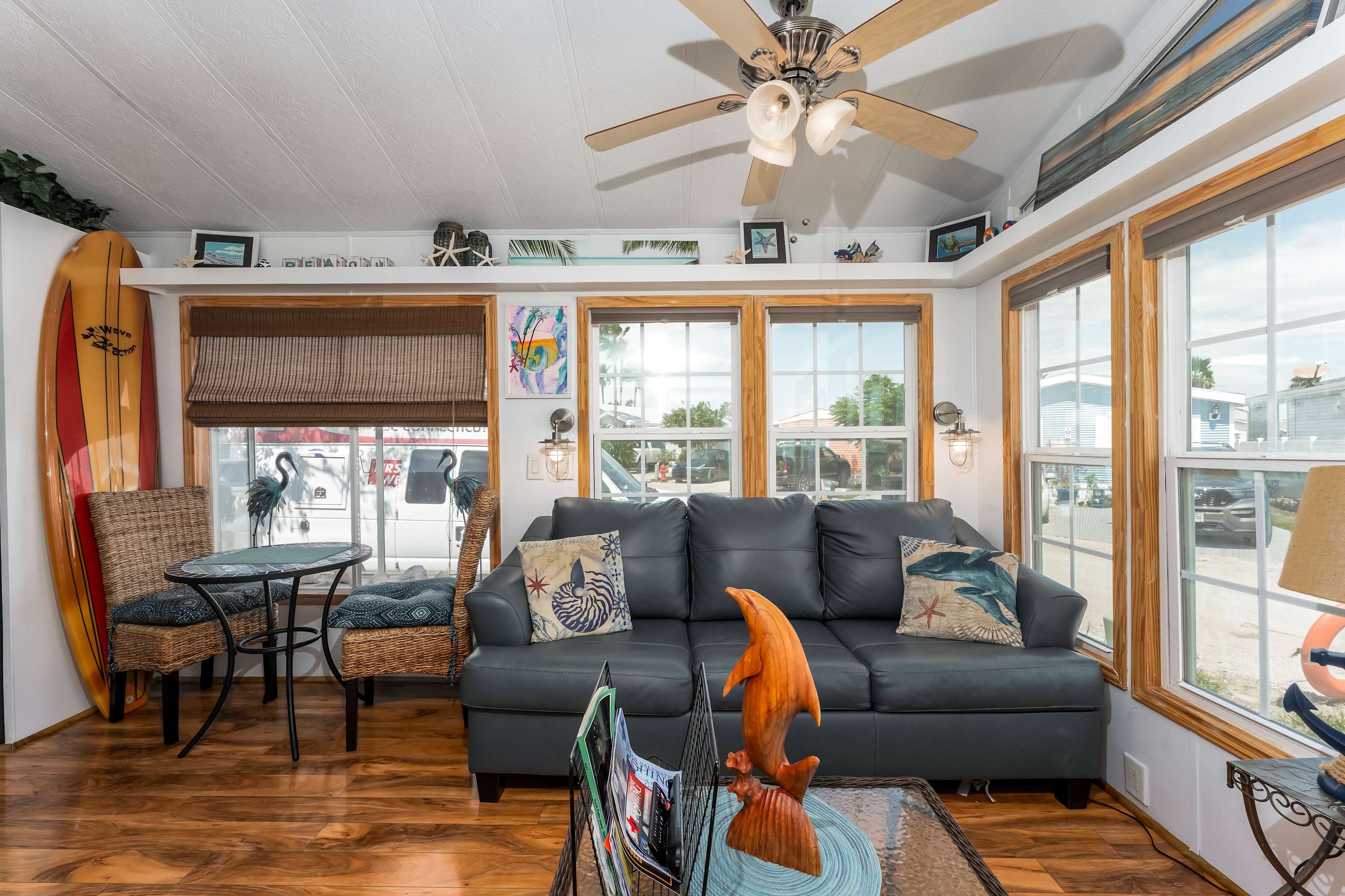 A cozy living room with a dark gray leather sofa, wood-look flooring, and multiple windows with wooden frames, decorated with coastal-themed accents, a ceiling fan, and a surfboard.