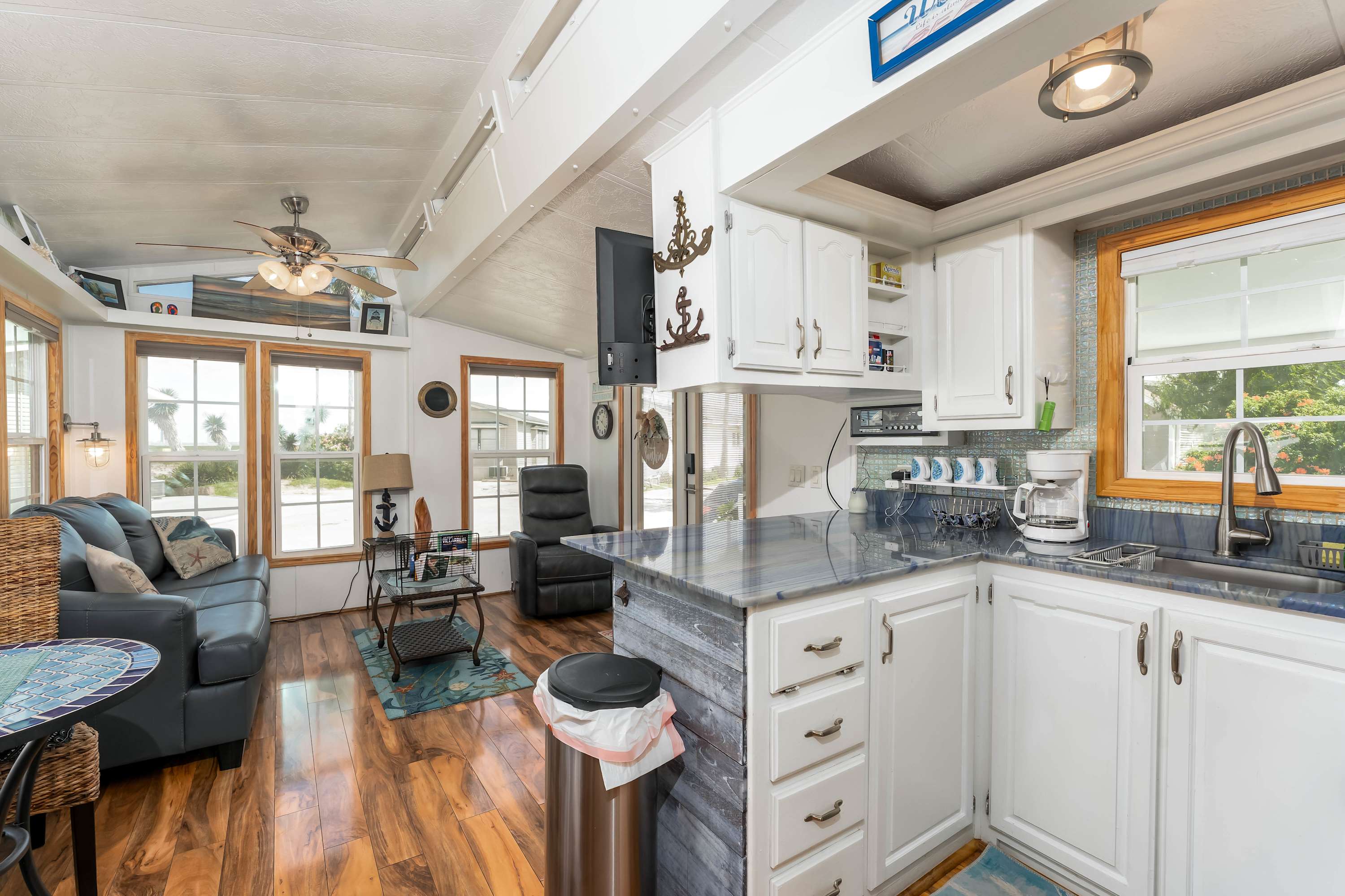 An open-concept living area and kitchen featuring wood-look flooring, a dark gray sofa, and a kitchen island with blue marble-patterned countertops, white cabinets, and a flat-screen TV mounted on a pillar.