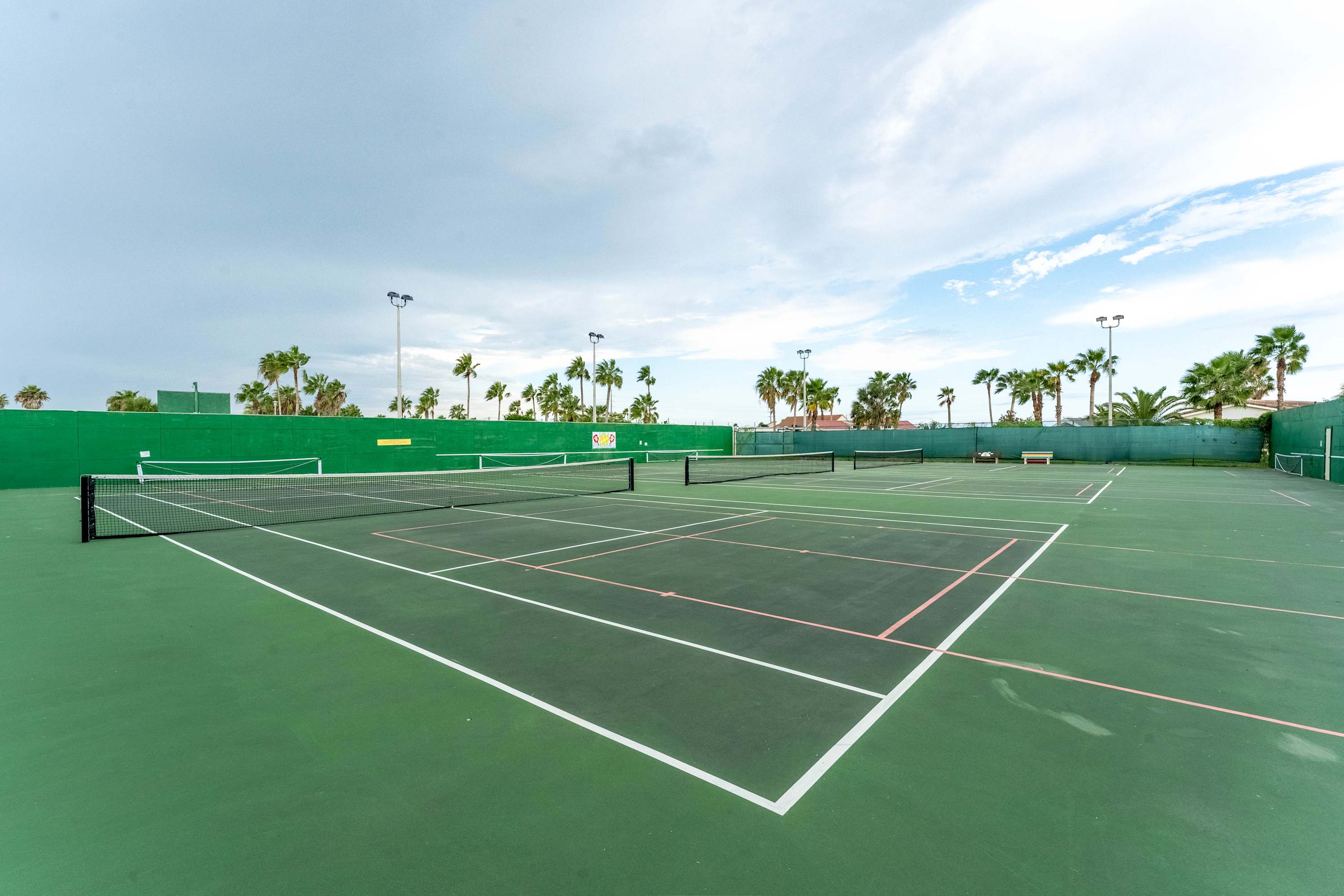 Multiple green tennis courts with white lines, surrounded by a tall green fence and numerous palm trees under a bright, partly cloudy sky. The courts appear well-maintained and ready for play.
