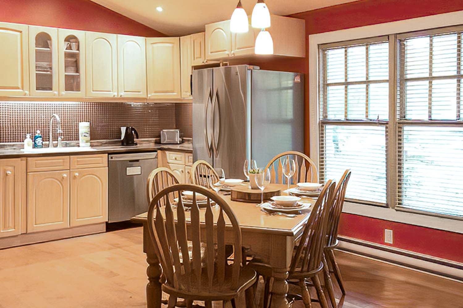 Well-lit kitchen and dining area with light wood cabinets, stainless steel refrigerator, and a wooden dining table set for six. The walls are a striking red, complemented by pendant lighting and a large window with blinds.