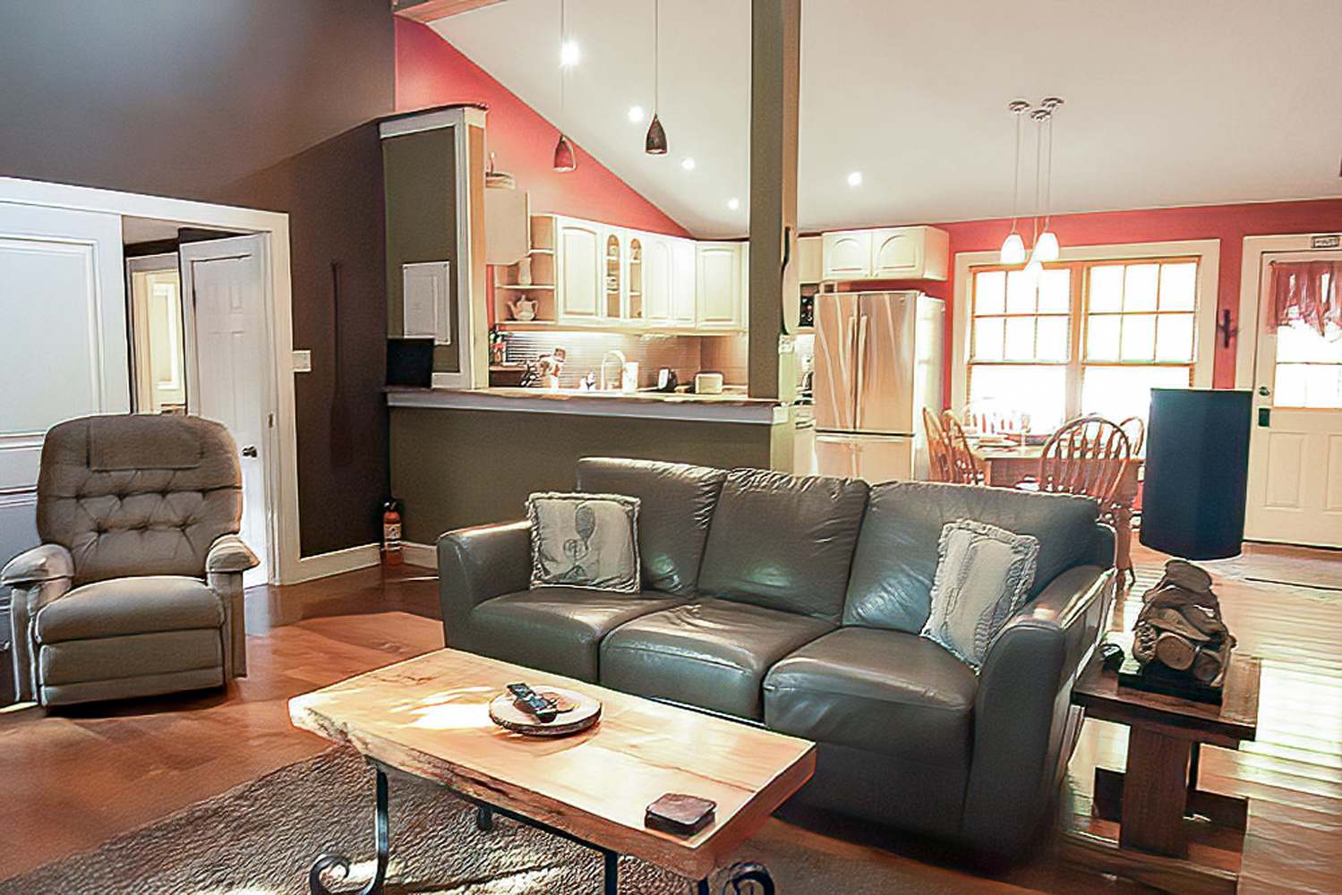 Open concept living area showing a brown leather sofa, a wooden coffee table, and a grey recliner, with a view into the kitchen and dining area. Hardwood floors extend throughout, and the walls are a mix of brown and red.