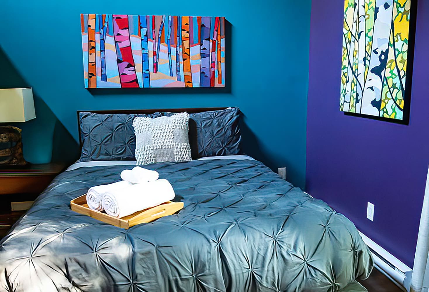Close-up of a queen bed with a gray tufted duvet and white rolled towels on a wooden tray, set against a vibrant teal and purple wall with abstract artwork.