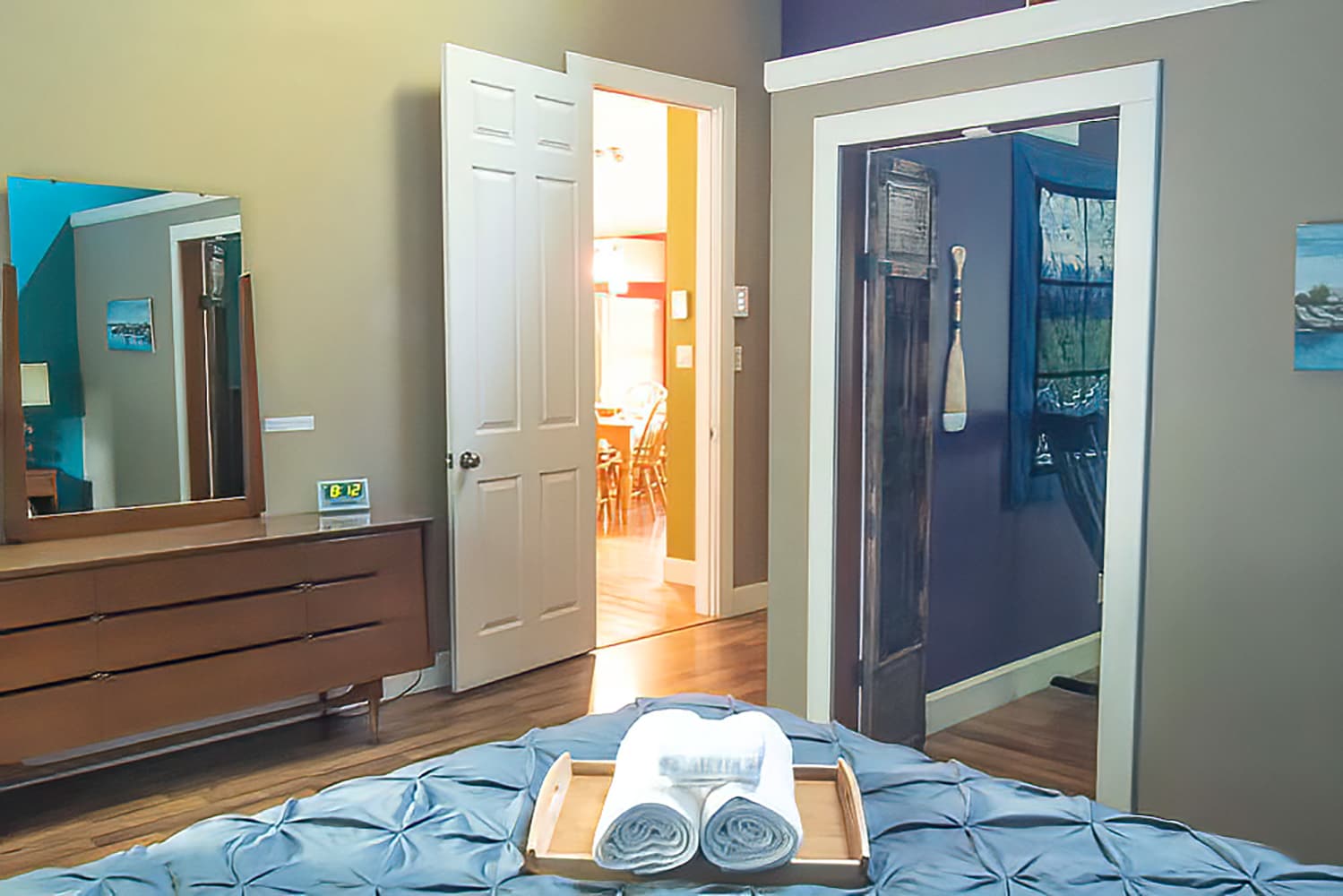 A bedroom with a blue tufted duvet and a wooden dresser, offering a view into an adjacent room through an open white door, with light brown walls.