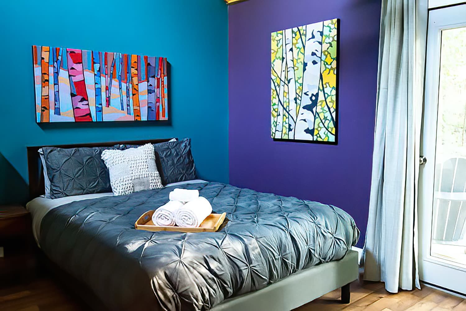 A vibrant bedroom featuring a queen bed with a gray tufted duvet, flanked by a teal accent wall and a purple wall, adorned with colorful abstract art.