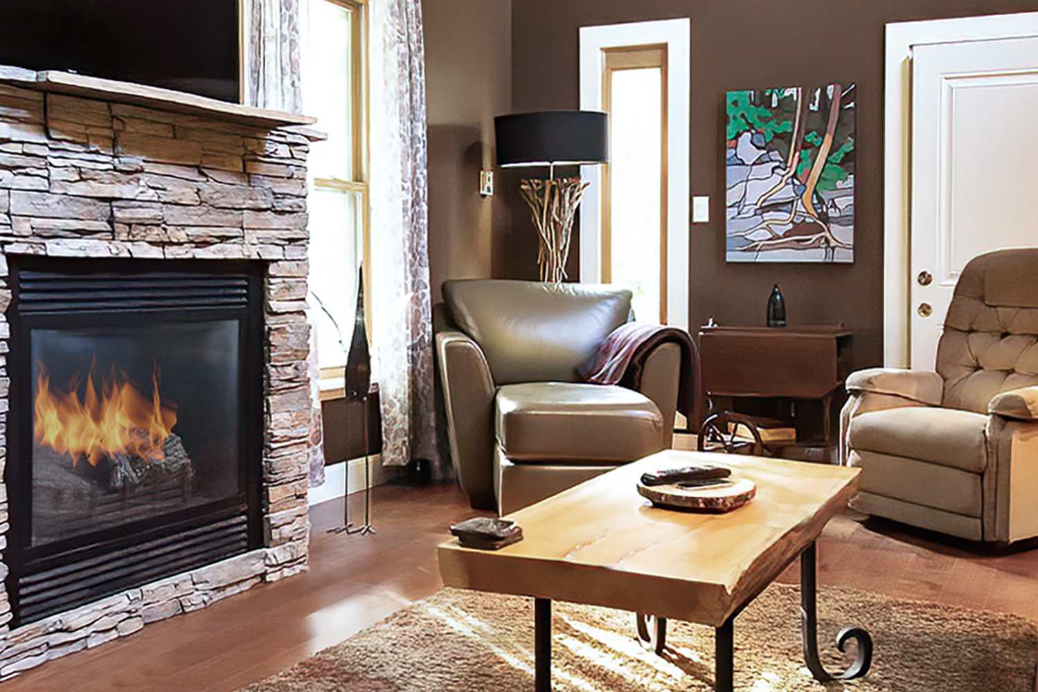 Cozy living room with a stone fireplace, a brown leather armchair, and a wooden coffee table on a shaggy rug. A large window with patterned curtains lets in natural light, and an abstract art piece hangs on the wall.