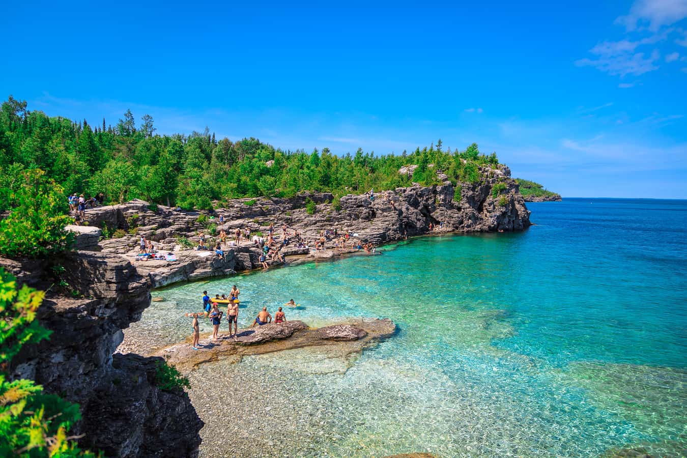 A vibrant natural cove featuring crystal-clear turquoise water, rugged rocky cliffs, and dense green forest under a bright blue sky. Many people are seen swimming in the water and relaxing on the sun-drenched rocks.