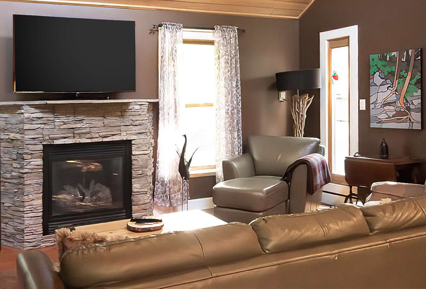 Inviting living room featuring a large flat-screen TV mounted above a stone fireplace, a brown leather sofa, and a leather armchair. Natural light streams through a window with sheer curtains, illuminating the hardwood floors.