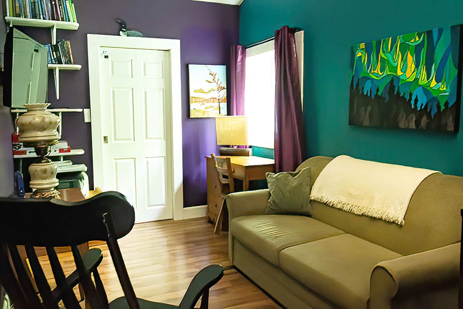 A vibrant living room with a teal accent wall and purple walls, featuring a light green sofa, a small wooden desk, wall-mounted shelves, and a dark rocking chair.