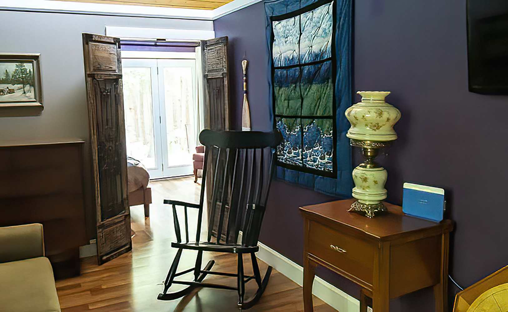 A cozy living area features a dark rocking chair, a wooden side table with a vintage lamp, and purple walls, with a glimpse into a bedroom through open doors.