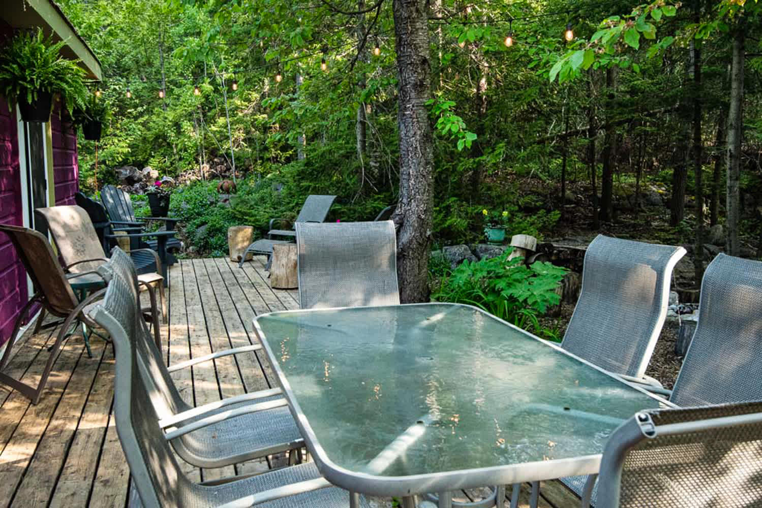 A spacious wooden deck nestled in a lush forest setting, featuring an outdoor dining table with chairs and string lights overhead, surrounded by trees and greenery.
