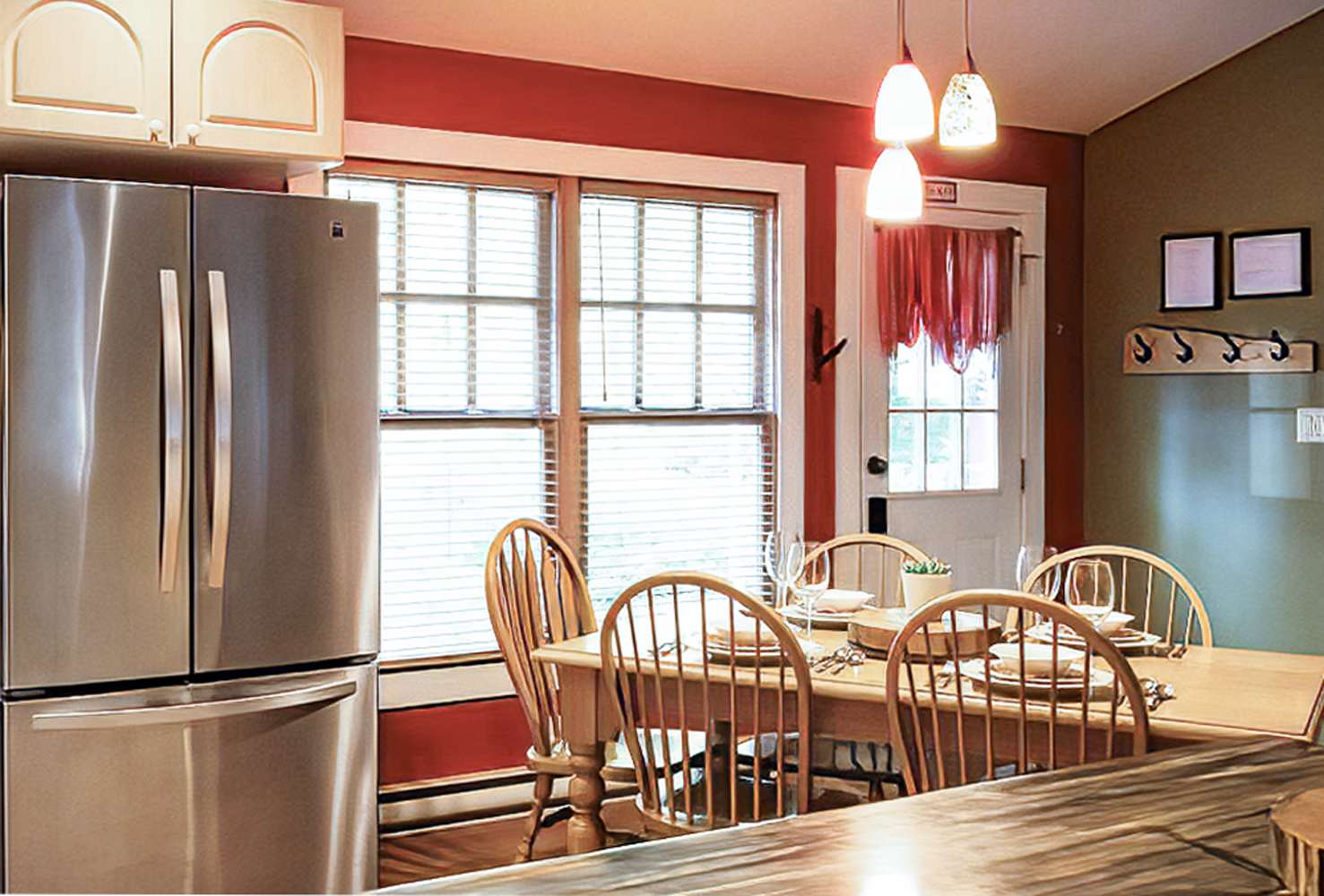 View of the kitchen and dining area, highlighting the stainless steel refrigerator, a wooden dining table set for six, and a large window with blinds. The red walls and pendant lights create a warm ambiance.