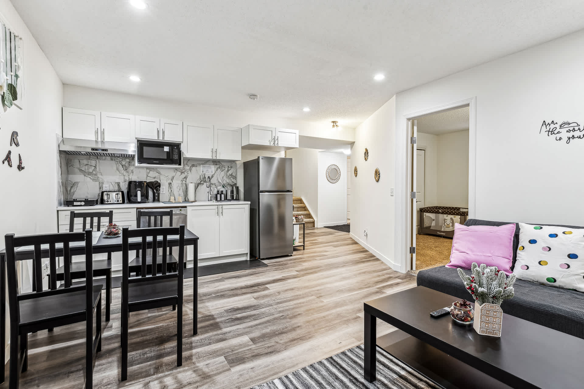 An open-concept living space with a dark grey sofa, a black coffee table, and a compact kitchen with white cabinets. Wood-look laminate flooring extends throughout, and a doorway leads to a carpeted bedroom.