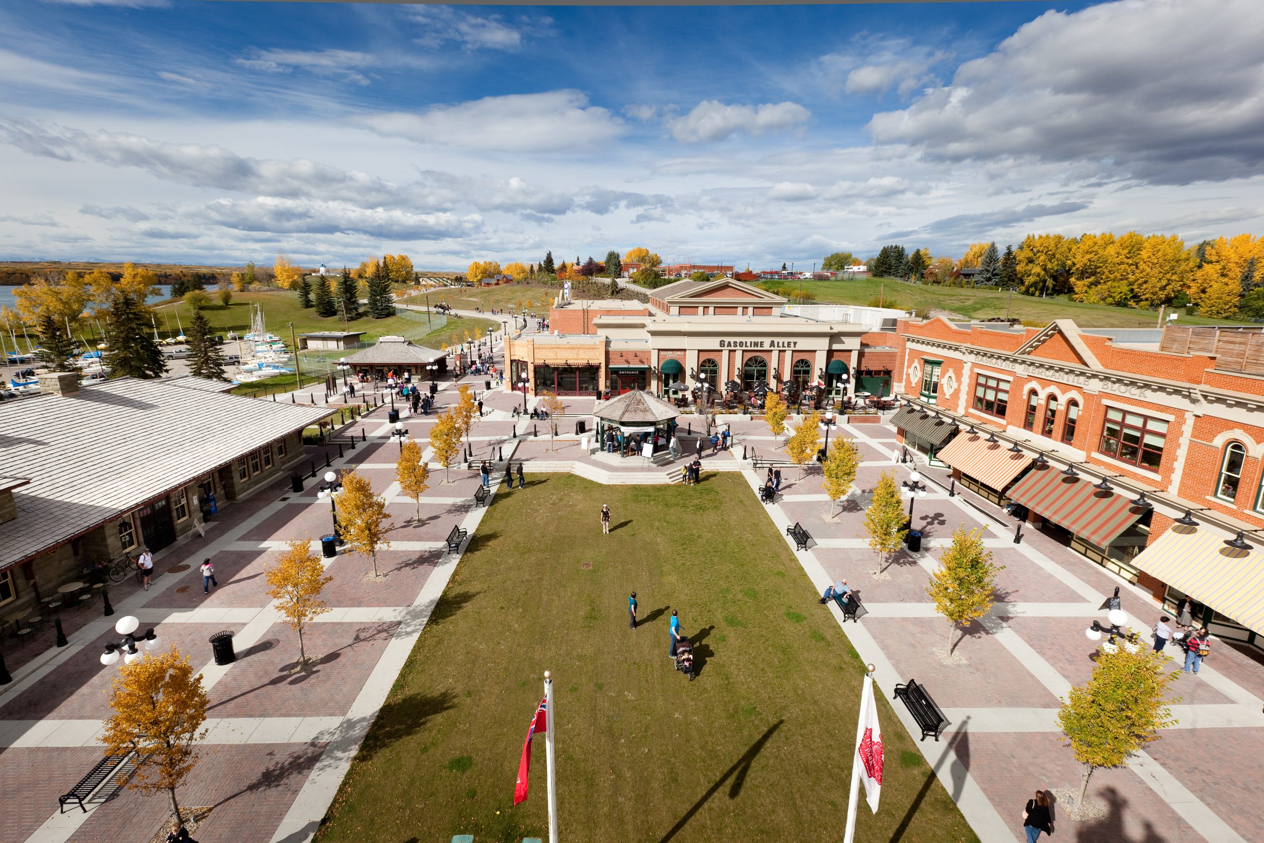 An aerial view of a historical village or park with numerous buildings, a large green lawn, and people strolling around, surrounded by trees with autumn foliage and a body of water in the background.