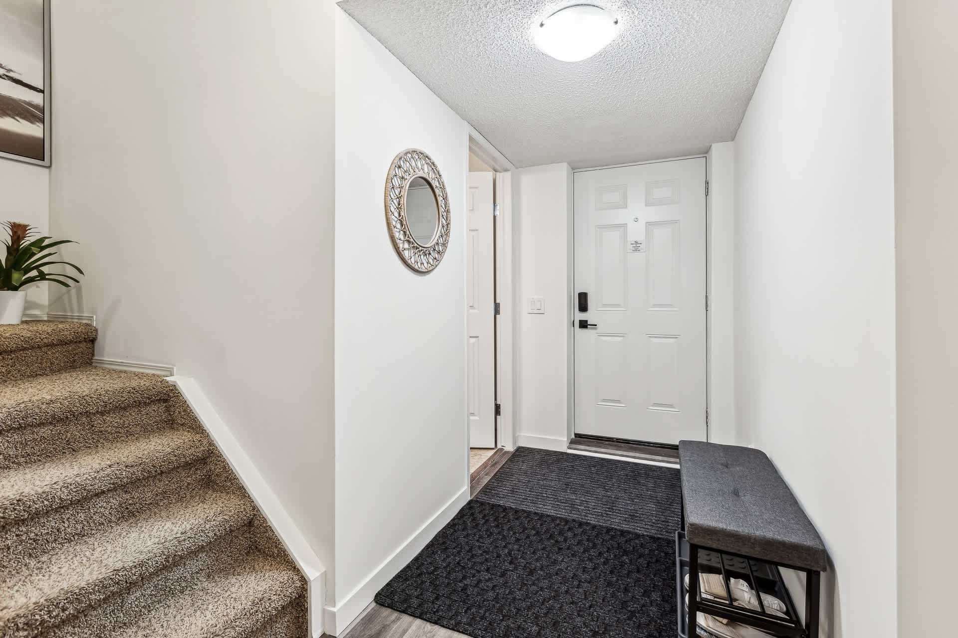 An entryway with dark wood-look laminate flooring, a black welcome mat, a white front door with a smart lock, and a small bench with shoe storage, leading to a carpeted staircase. A round decorative mirror hangs on the wall.