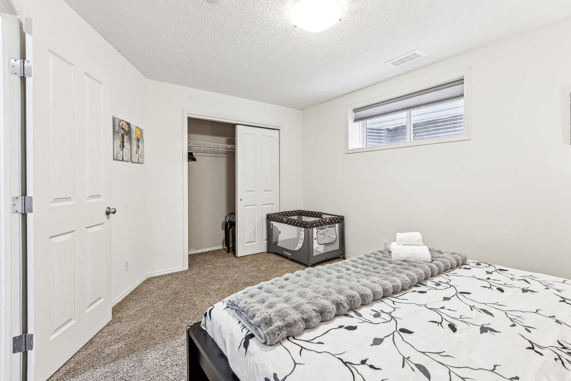 A bright bedroom with light brown carpet, featuring a dark wood bed frame with a patterned comforter. A closet with bi-fold doors is visible, along with a portable crib (pack-n-play) in the corner, indicating child-friendly amenities.