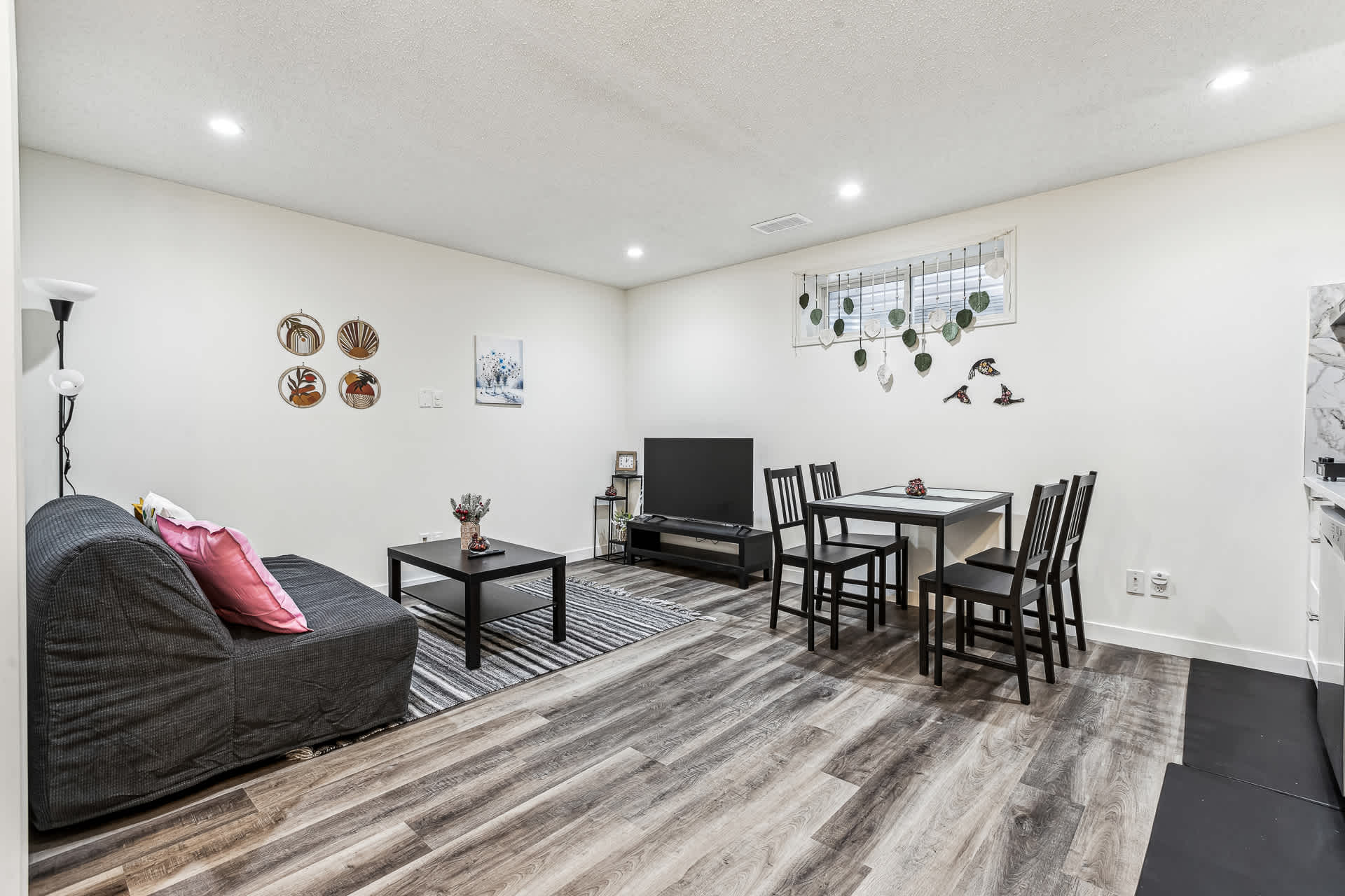 A spacious open-concept living and dining area with wood-look laminate flooring and white walls. A dark grey sofa and coffee table define the living space, while a black dining table with four chairs is positioned near a window.