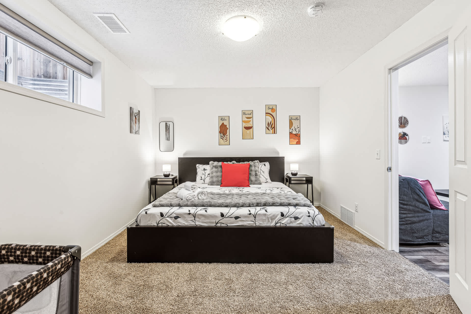 A well-lit bedroom featuring a dark wood bed frame with a patterned comforter and a red accent pillow. Black nightstands with small lamps flank the bed, and abstract art decorates the white wall above the headboard.