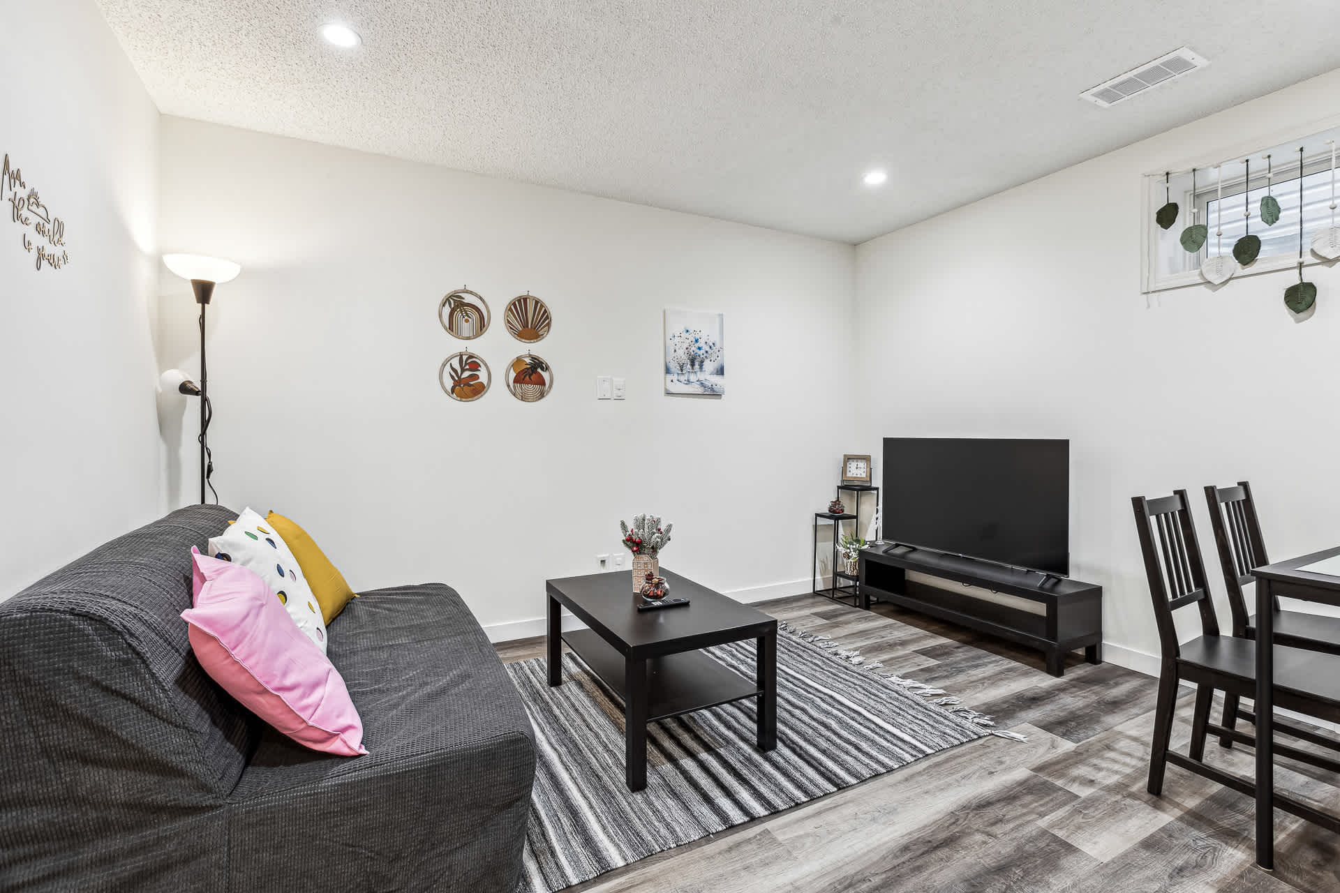 A bright living room featuring a dark grey sofa with colorful pillows, a black coffee table on a striped rug, and a wall-mounted TV. A floor lamp stands in the corner, and decorative wall art adorns the white walls.