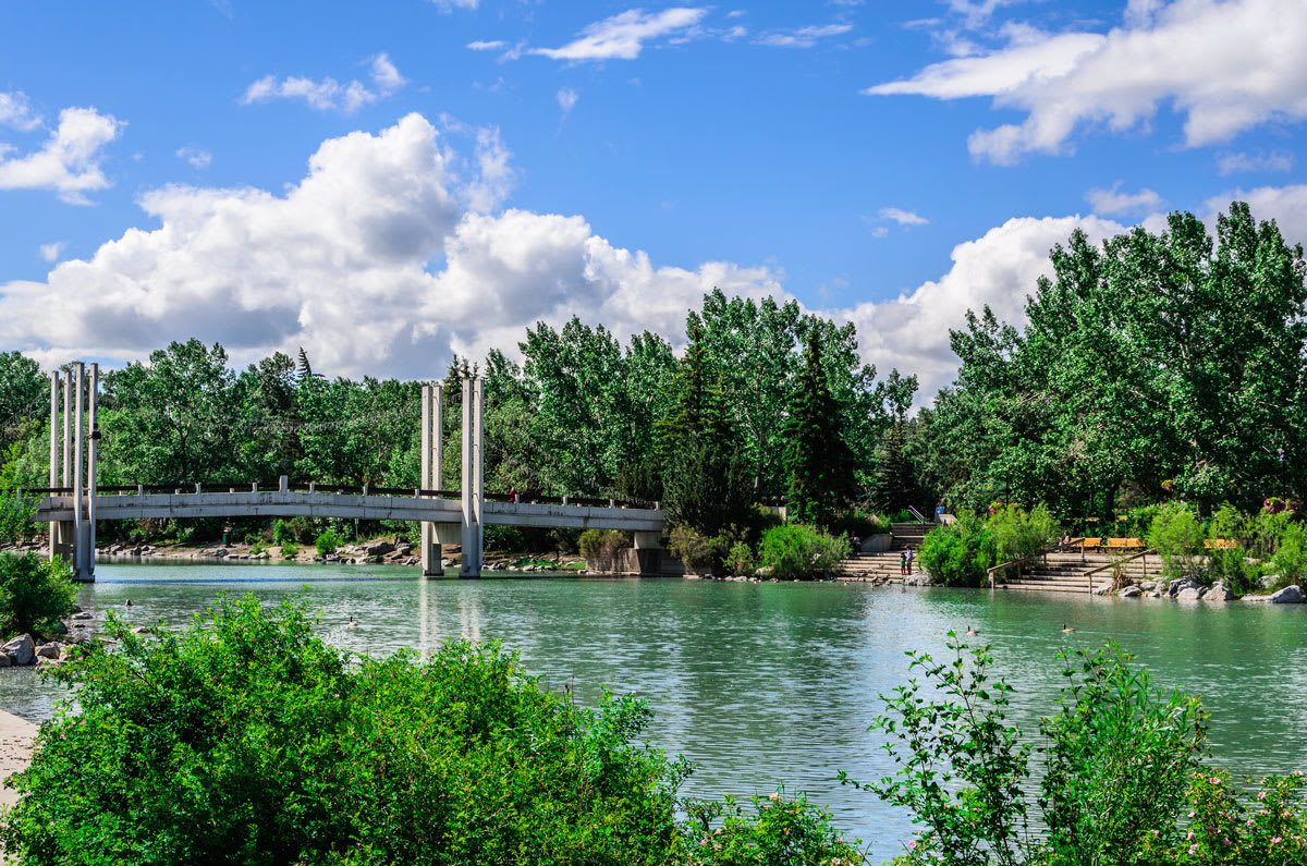 A wide shot of a serene river with a modern pedestrian bridge spanning across it, surrounded by dense green trees and a clear blue sky with fluffy white clouds.