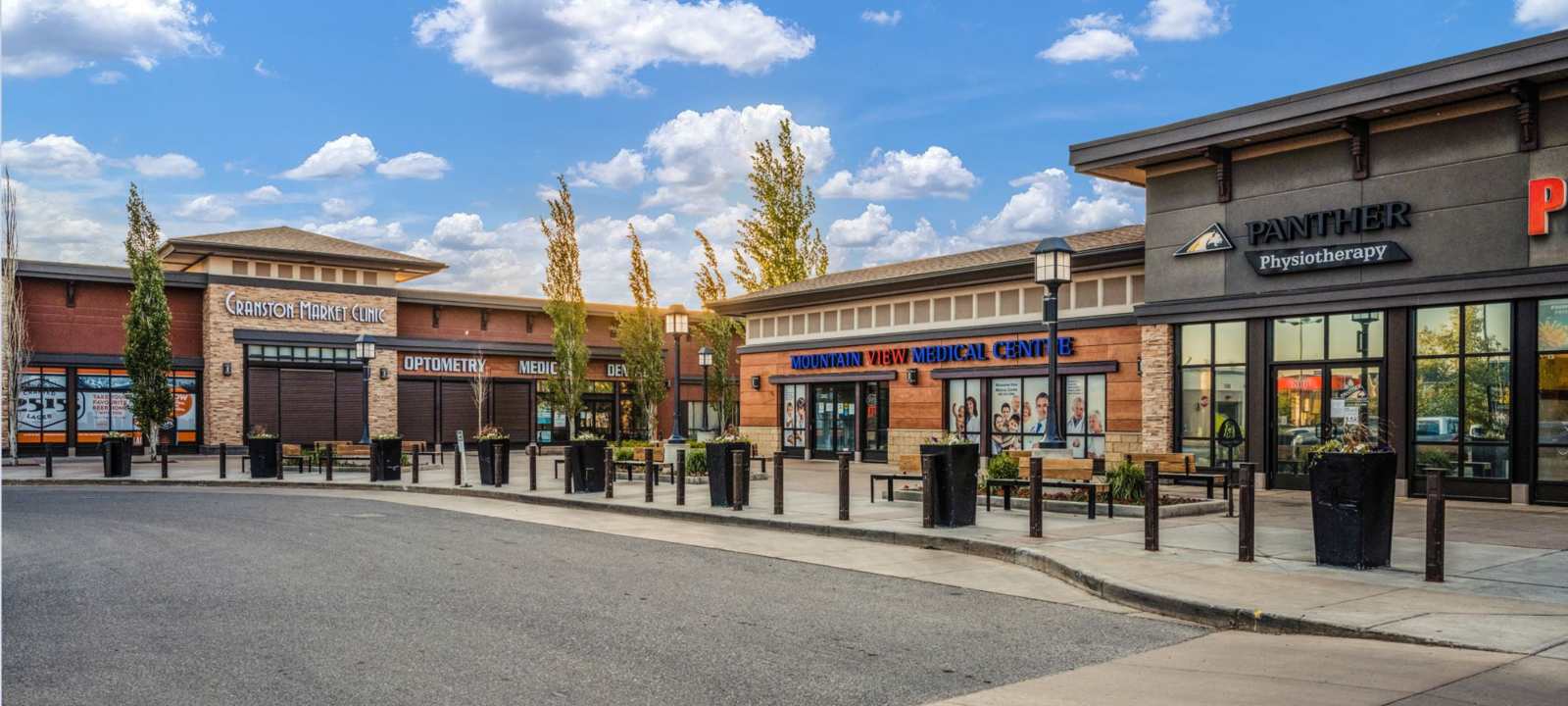 An outdoor view of a modern commercial plaza with various businesses including a medical center, optometry, and physiotherapy, featuring brick facades, large windows, and outdoor seating areas under a partly cloudy sky.