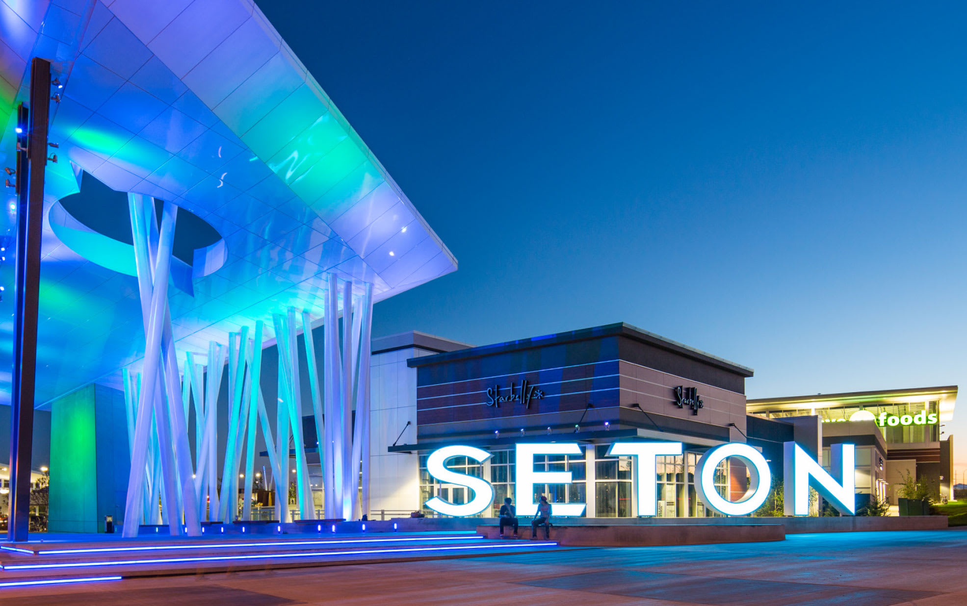 A vibrant night view of the Seton shopping district, highlighted by a large illuminated 'SETON' sign and modern buildings with colorful blue and green lighting.